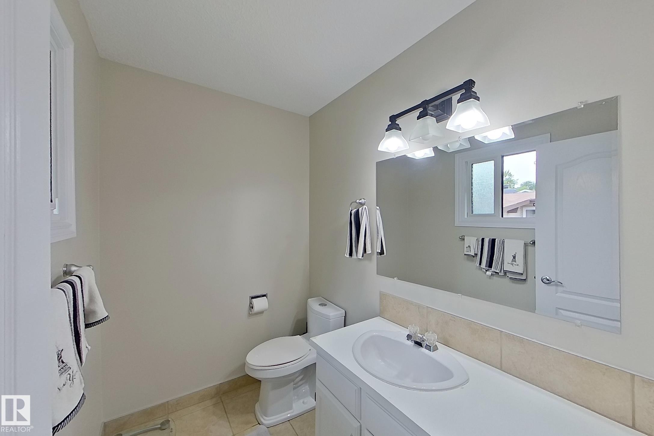Bright bathroom featuring dual vanity with white cabinetry and wood-finish countertop, large framed mirror with integrated lighting, ceramic tile flooring, and updated fixtures throughout - 14 Brunswick Crescent, St. Albert, AB