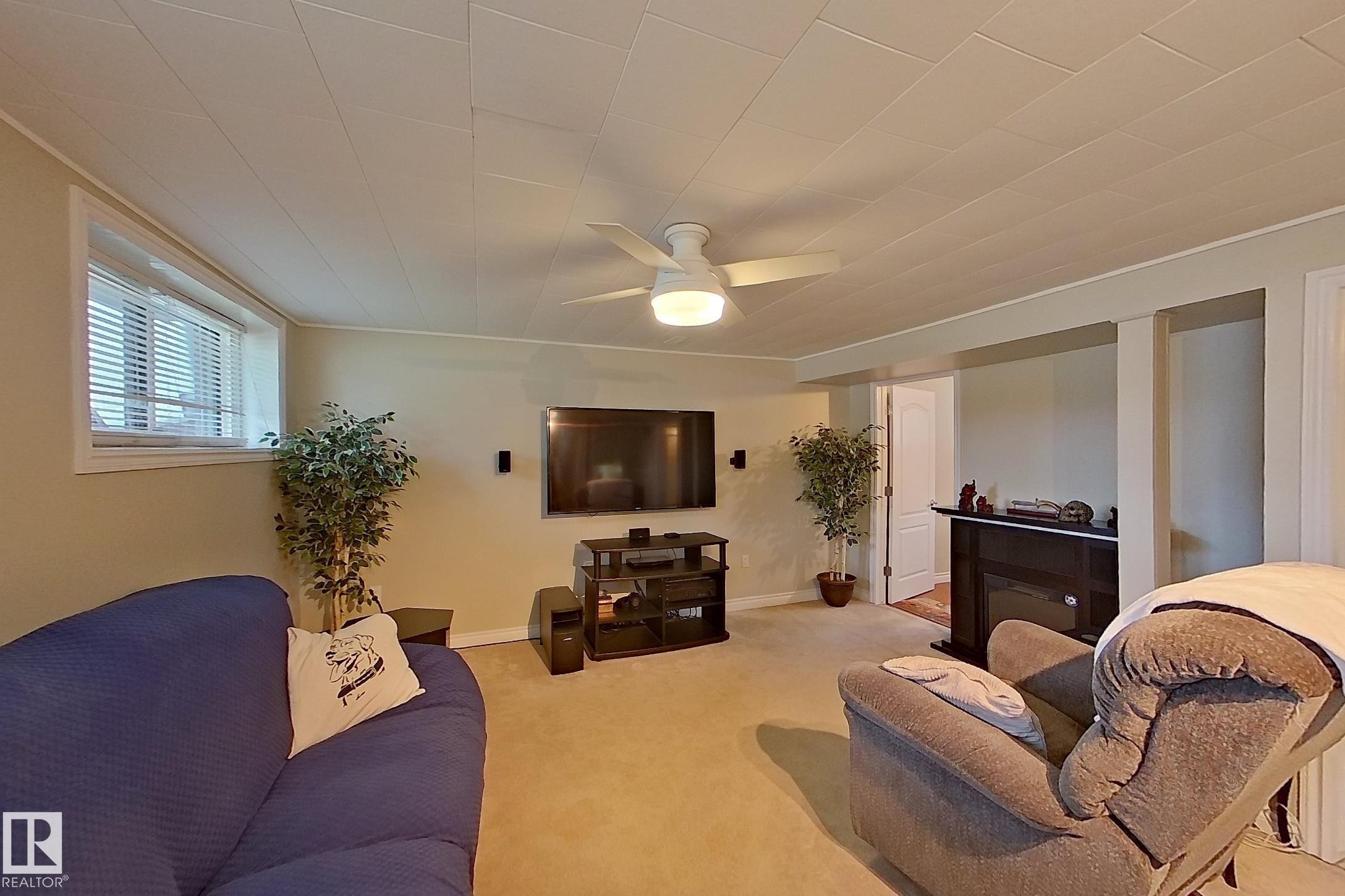 Spacious recreation room featuring neutral carpeting, recessed ceiling lighting with ceiling fan, wall-mounted flat-screen television, built-in shelving with dark wood finish, and multiple windows providing natural light - 14 Brunswick Crescent, St. Albert, AB