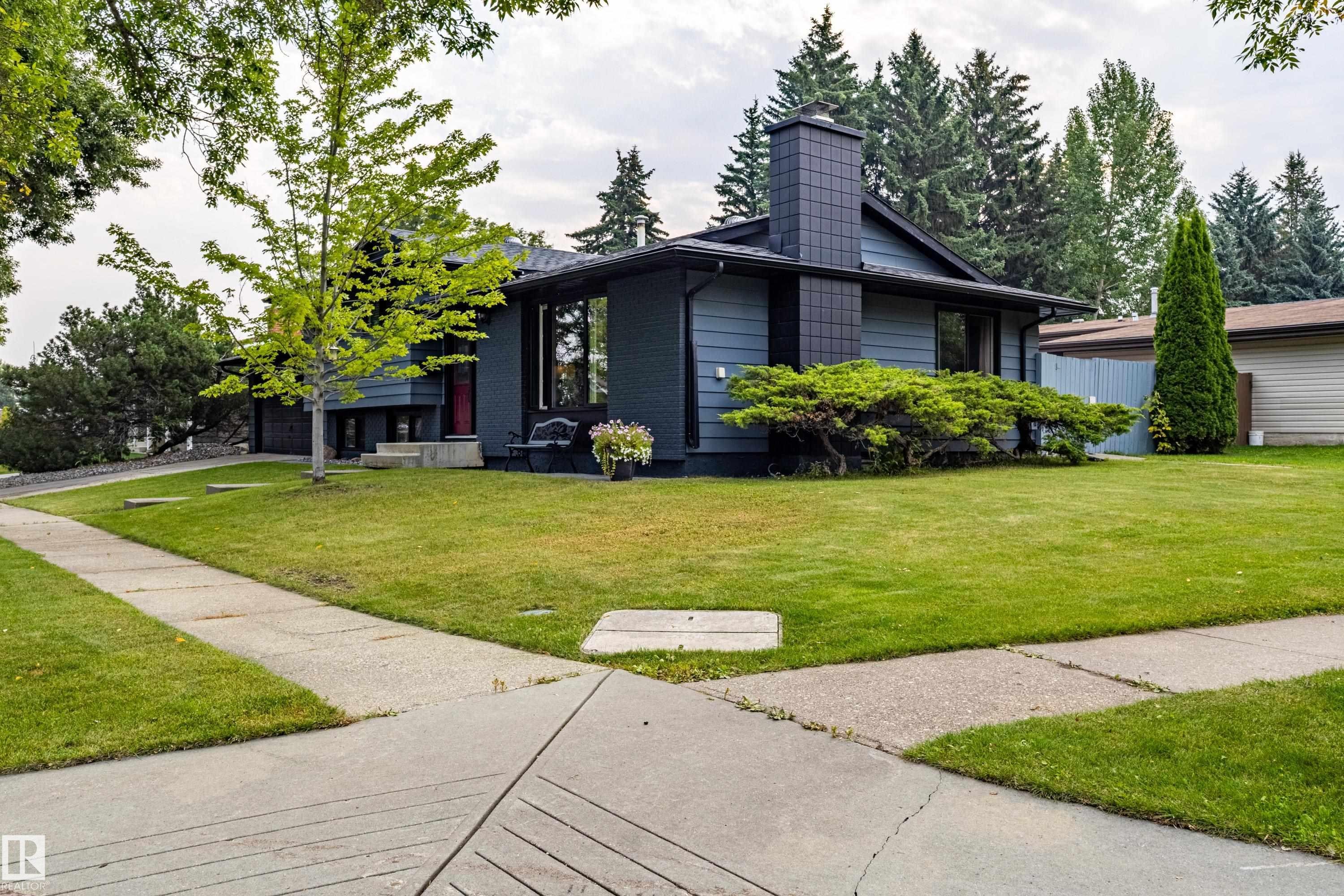 Contemporary home featuring a modern black exterior with geometric architectural lines, expansive manicured lawn with mature landscaping, concrete driveway with clean sightlines, and abundant natural tree coverage throughout the property - 14 Brunswick Crescent, St. Albert, AB