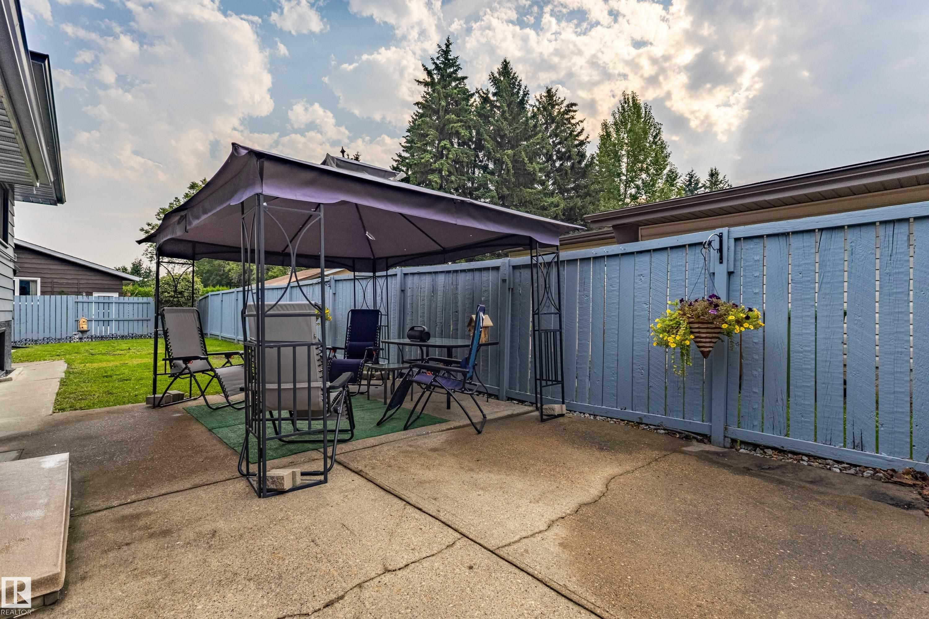 Outdoor entertaining space featuring a retractable canopy shade structure, concrete patio foundation, and mature landscaping with evergreen trees - 14 Brunswick Crescent, St. Albert, AB