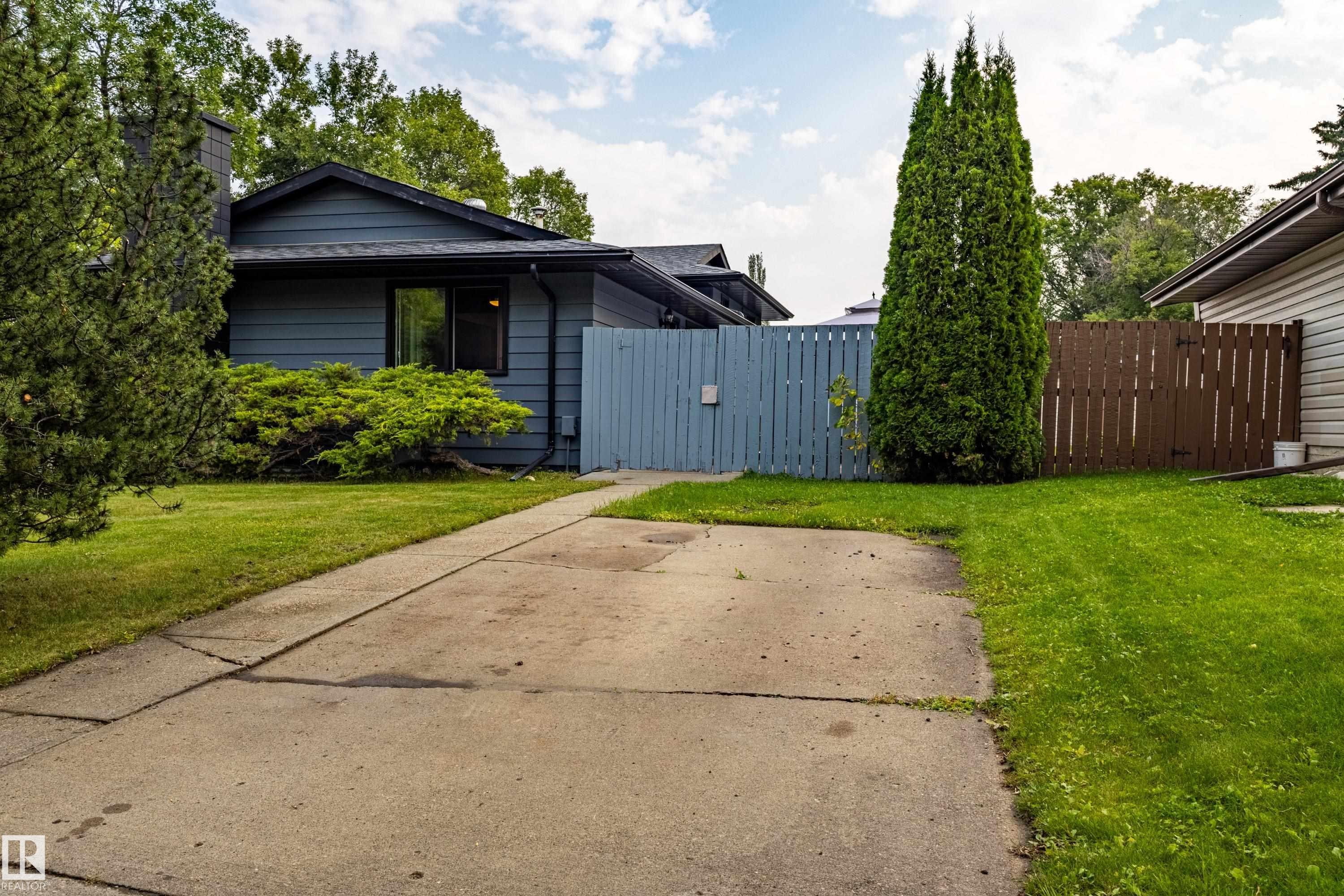 Welcoming single-story residence with a spacious concrete driveway, mature landscaping including tall evergreen specimens, and lush lawn areas flanking the home - 14 Brunswick Crescent, St. Albert, AB