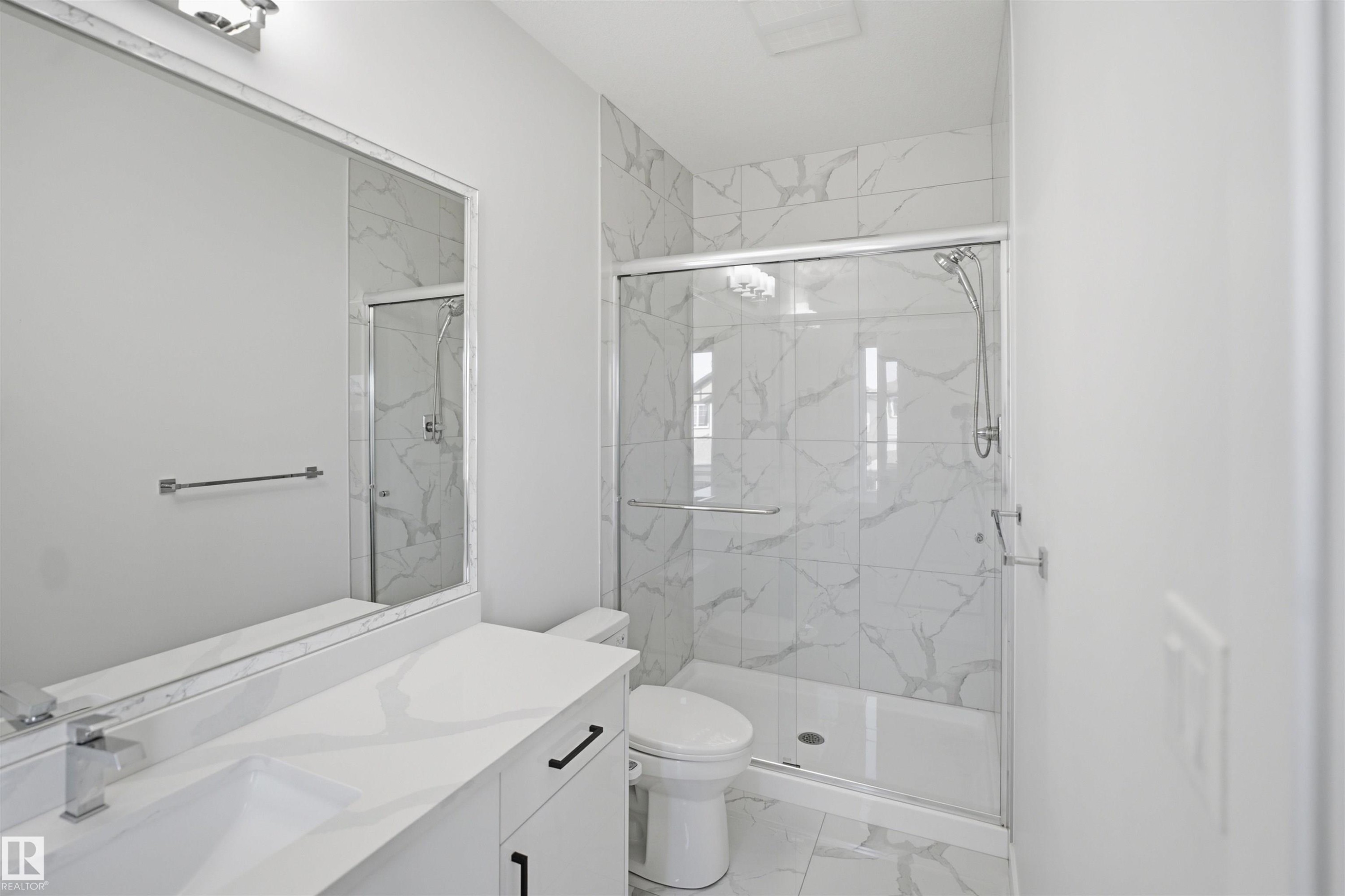 Vanity with integrated rectangular sink and chrome faucet, full-width wall mirror with overhead lighting, glass-enclosed shower with tiled surround, and white toilet - 2027 13 Avenue Nw, Edmonton, AB