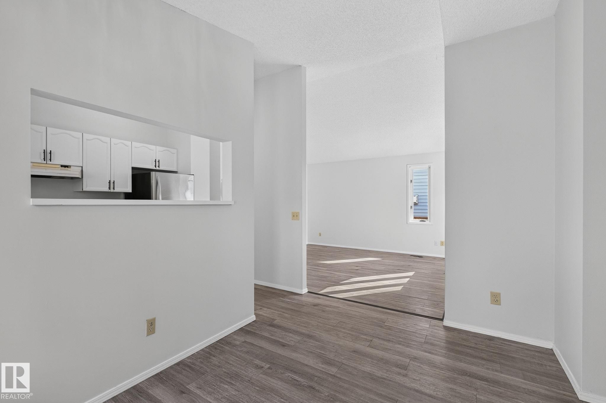 Open concept interior featuring wood-finish flooring, white walls, and a pass-through window to the kitchen with white cabinetry and a stainless steel refrigerator - 3722 34A Avenue Nw, Edmonton, AB - Indoor Photo Showing Other Room