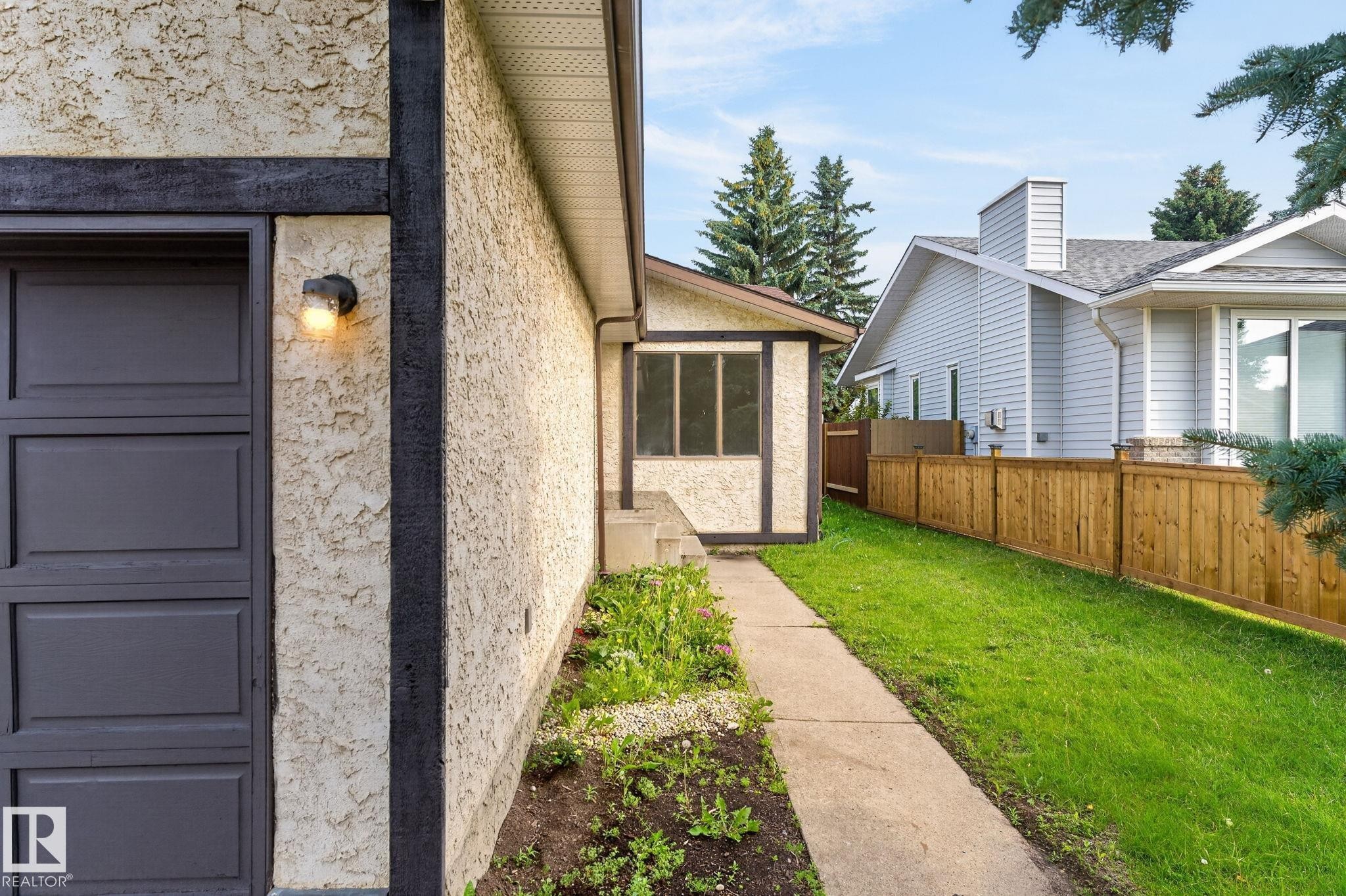 Stucco exterior with dark trim accents, featuring a side-entry garage door, an outdoor light fixture, a concrete walkway, and a fenced yard with established greenery - 3722 34A Avenue Nw, Edmonton, AB - Outdoor