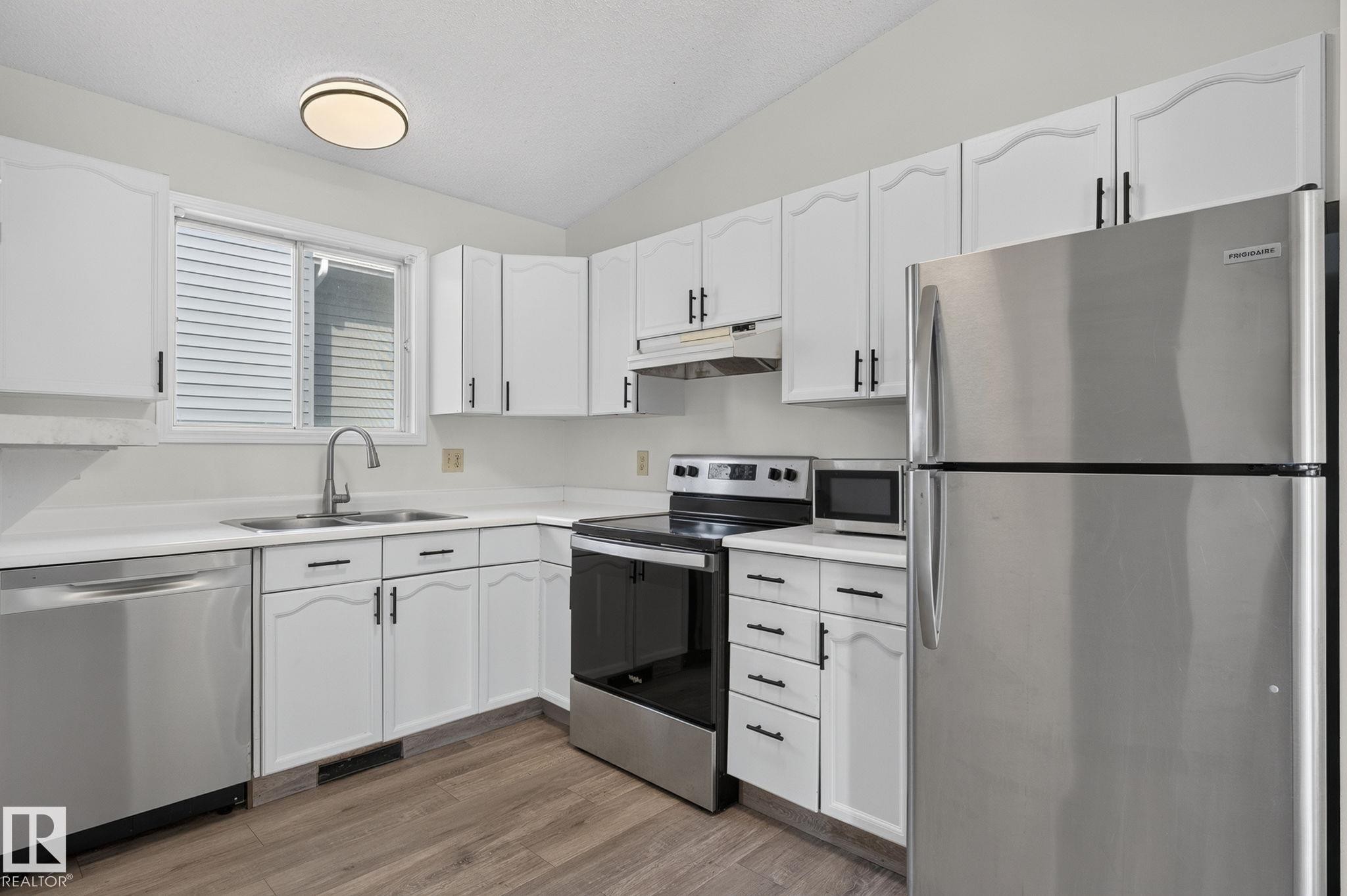 Kitchen featuring wood-finish flooring, white cabinetry with black hardware, and stainless steel appliances - 3722 34A Avenue Nw, Edmonton, AB - Indoor Photo Showing Kitchen With Double Sink