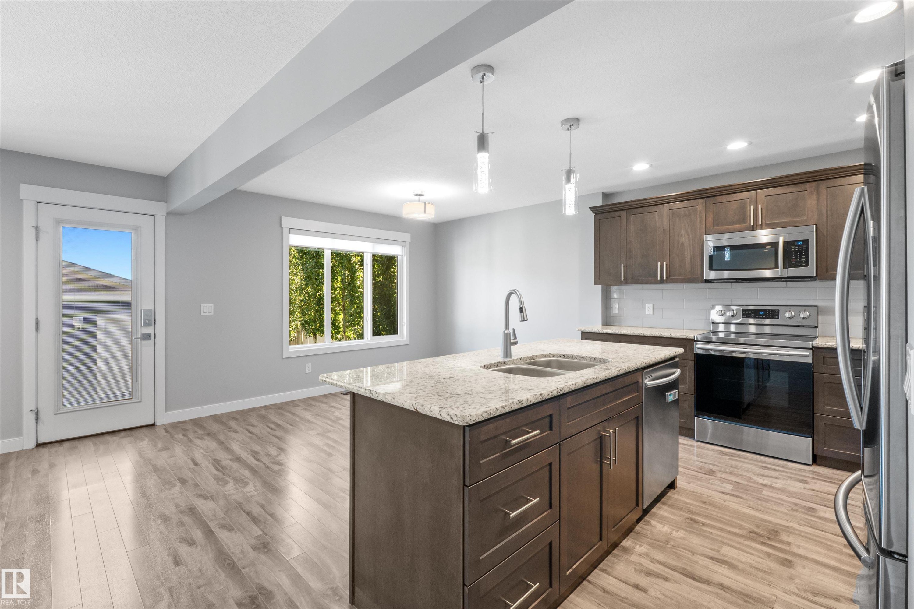 Open-concept kitchen featuring an island with a double basin sink, stainless steel appliances, wood-finish flooring, recessed lighting, and a glass-paneled exterior door - 2585 Pegasus Boulevard Nw, Edmonton, AB