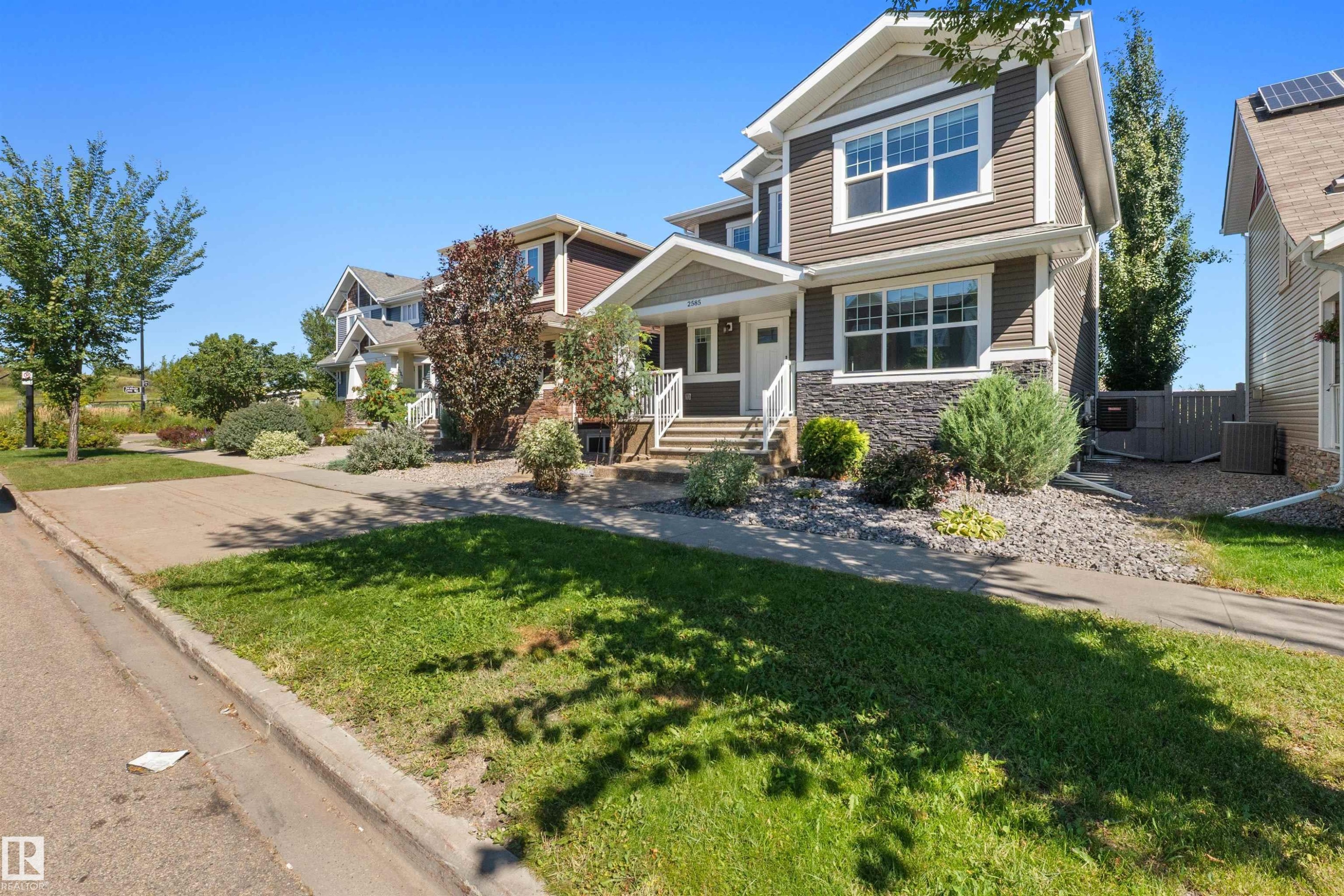 Two-story home with dark siding and stone-finish accents, featuring a front porch with white railings, a landscaped front yard with shrubs and gravel, and a concrete driveway - 2585 Pegasus Boulevard Nw, Edmonton, AB