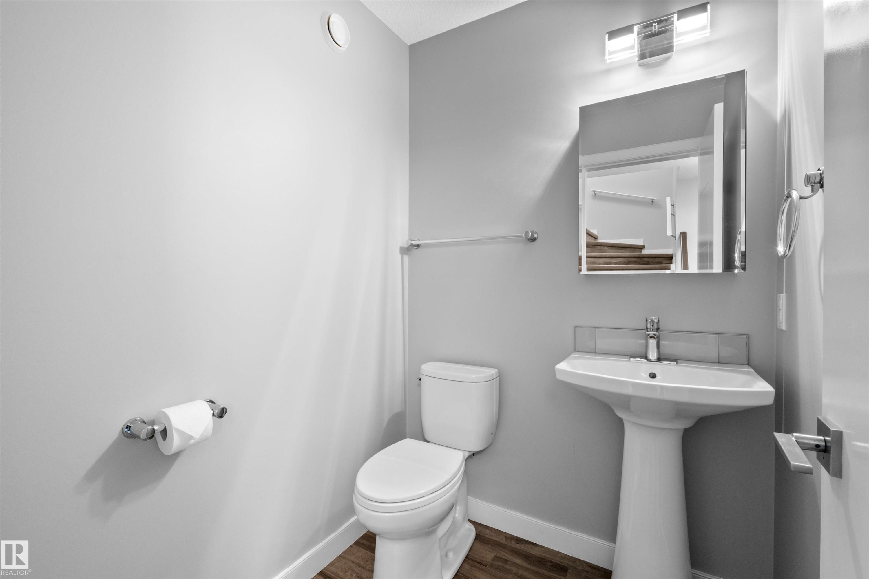 Pedestal sink with chrome faucet, wall-mounted mirror, overhead vanity lighting, white toilet, and wood-finish flooring - 2585 Pegasus Boulevard Nw, Edmonton, AB