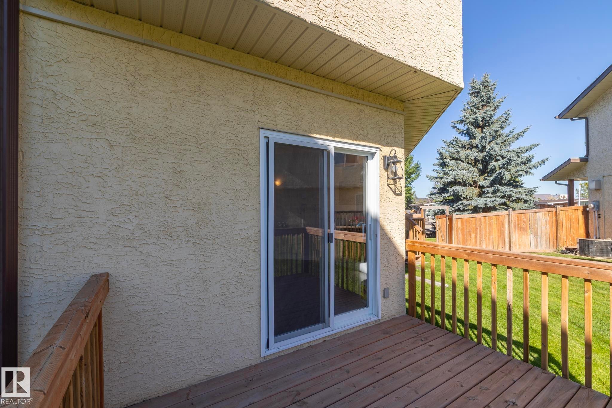 Wood deck with railing, sliding glass door, exterior wall lantern, stucco exterior, and fenced yard - #37 3811 85 St Nw, Edmonton, AB