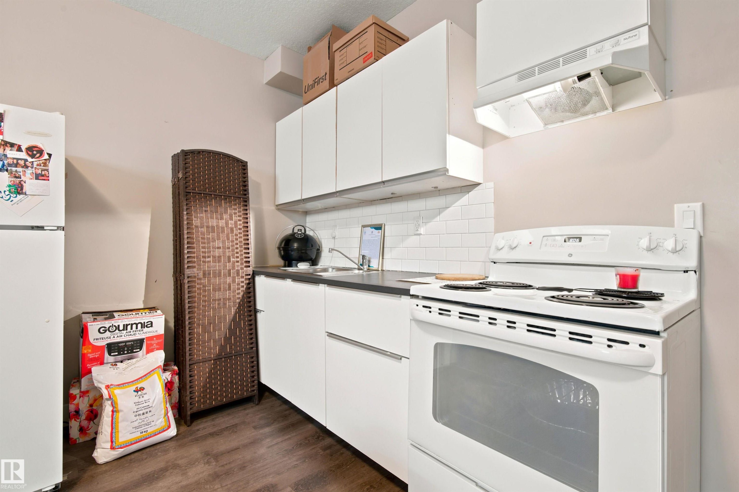 White cabinetry with modern hardware, dark countertops, white subway tile backsplash, stainless steel sink, and wood-finish flooring - 10833 63 Avenue Nw, Edmonton, AB