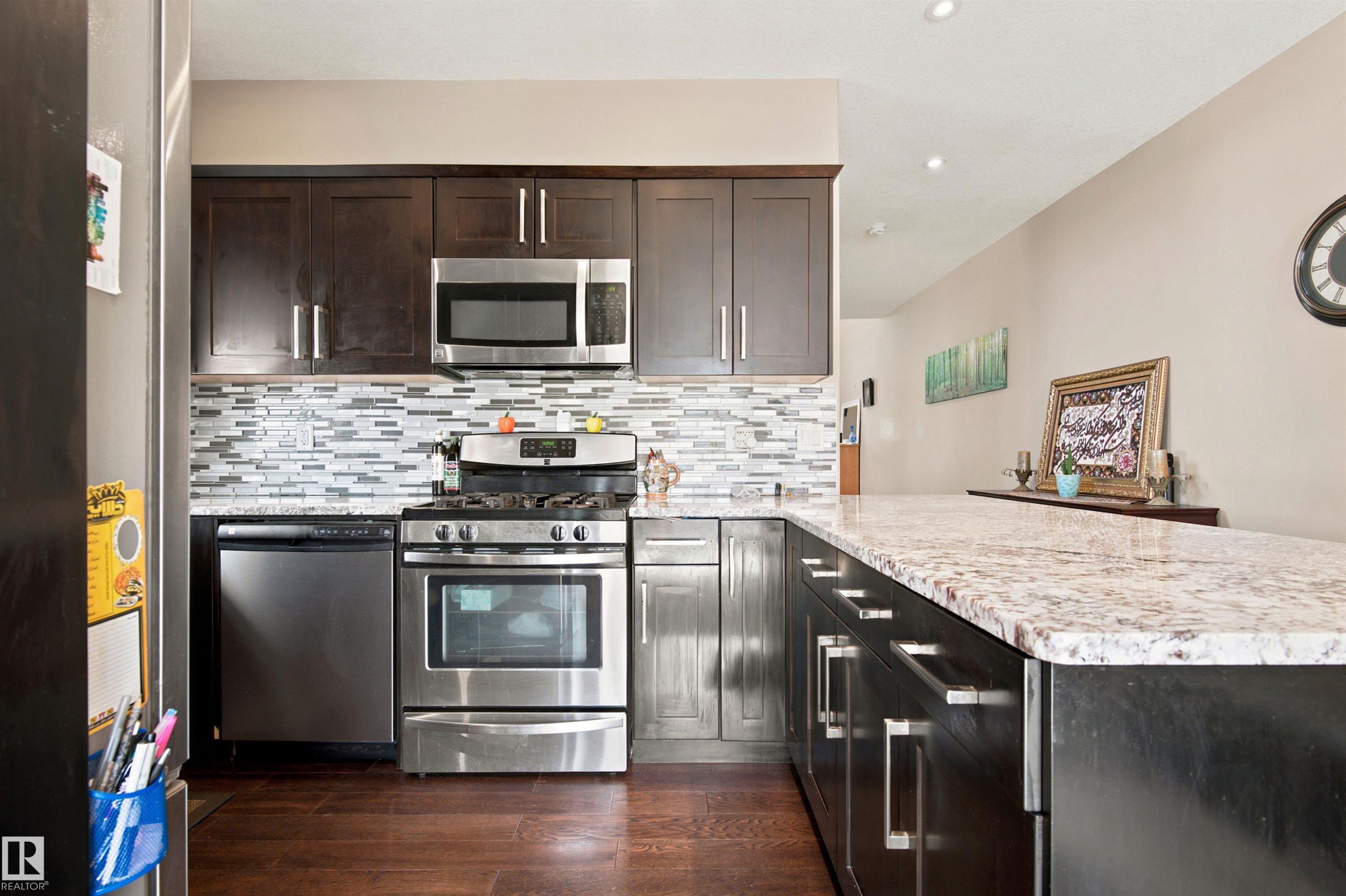 Kitchen featuring stainless steel appliances, dark wood-finish cabinetry, a patterned tile backsplash, light-colored stone countertops, and recessed lighting - 10833 63 Avenue Nw, Edmonton, AB