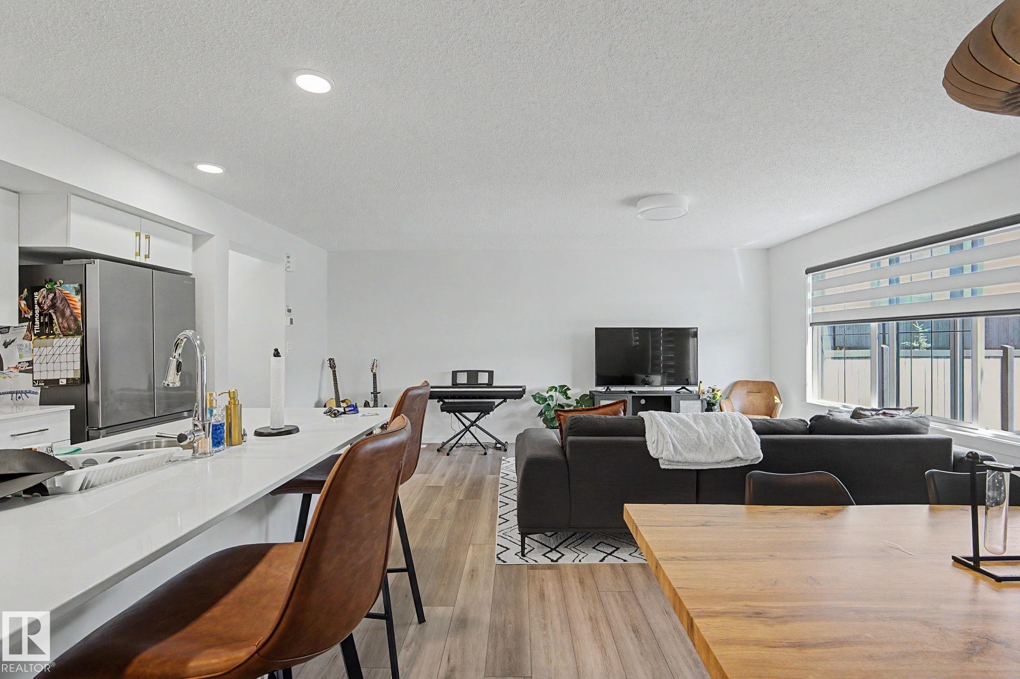 Open-concept living area featuring wood-finish flooring, recessed lighting, white cabinetry with gold-tone hardware, a stainless steel refrigerator, and a kitchen island with an integrated sink - #12 905 172 St Sw, Edmonton, AB - Indoor