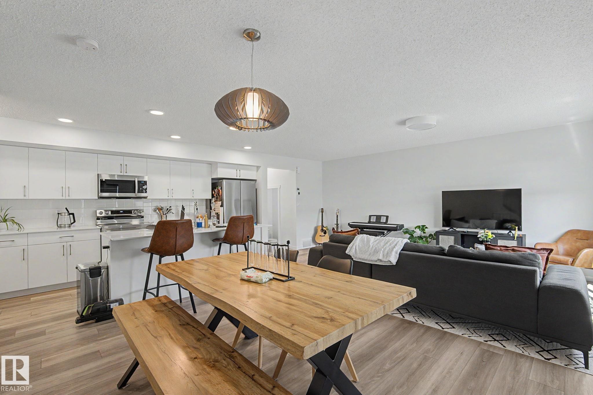 Open-concept living area featuring wood-finish flooring, a contemporary kitchen with white cabinetry, stainless steel appliances, a breakfast bar, and a dining area with a distinctive overhead light fixture - #12 905 172 St Sw, Edmonton, AB - Indoor