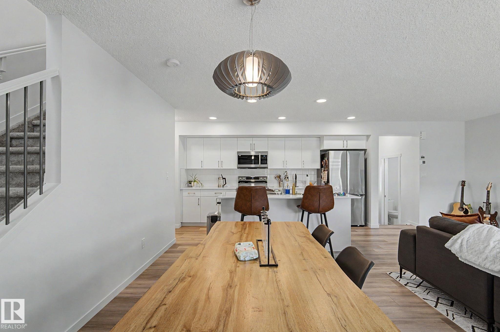 Open-concept living space featuring wood-finish flooring throughout, a modern pendant light fixture, and a kitchen with white cabinetry and stainless steel appliances - #12 905 172 St Sw, Edmonton, AB - Indoor