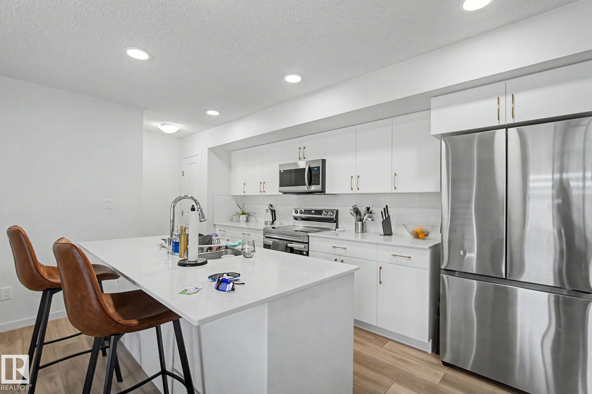 Modern kitchen featuring a white island with an integrated sink, stainless steel appliances, white cabinetry with gold-tone hardware, and wood-finish flooring - #12 905 172 St Sw, Edmonton, AB - Indoor Photo Showing Kitchen With Upgraded Kitchen