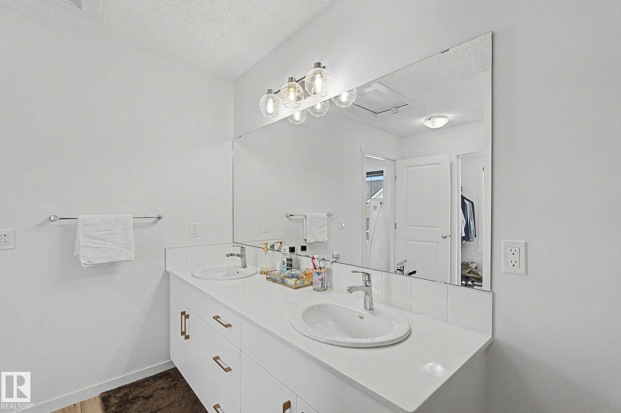 White dual vanity with integrated sinks and brass-finish hardware, illuminated by a contemporary light fixture, set against a full-width mirror - #12 905 172 St Sw, Edmonton, AB - Indoor Photo Showing Bathroom