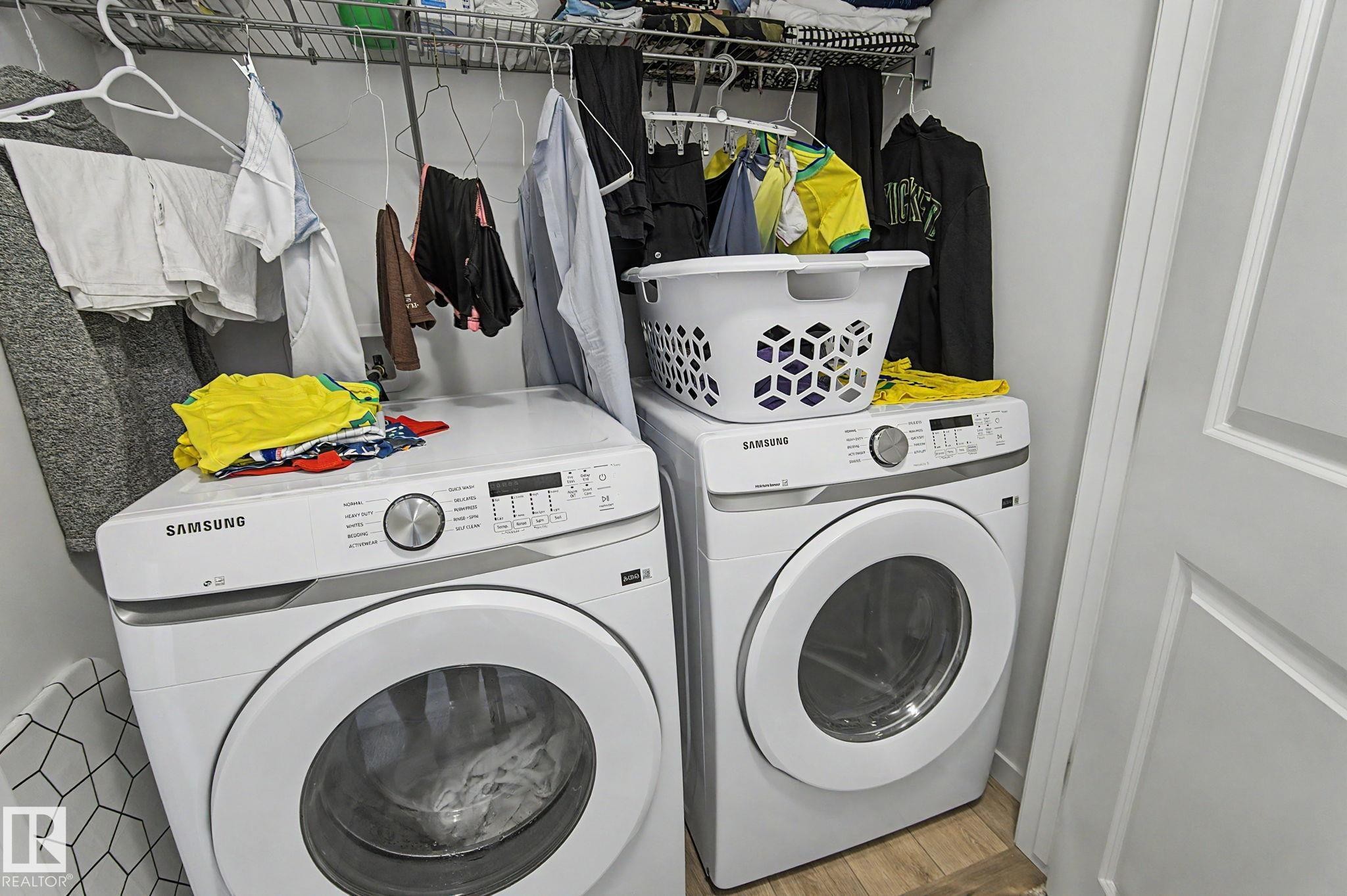 Dedicated laundry area featuring a white front-loading washer and dryer set, a wire shelving system, and wood-finish flooring - #12 905 172 St Sw, Edmonton, AB - Indoor Photo Showing Laundry Room