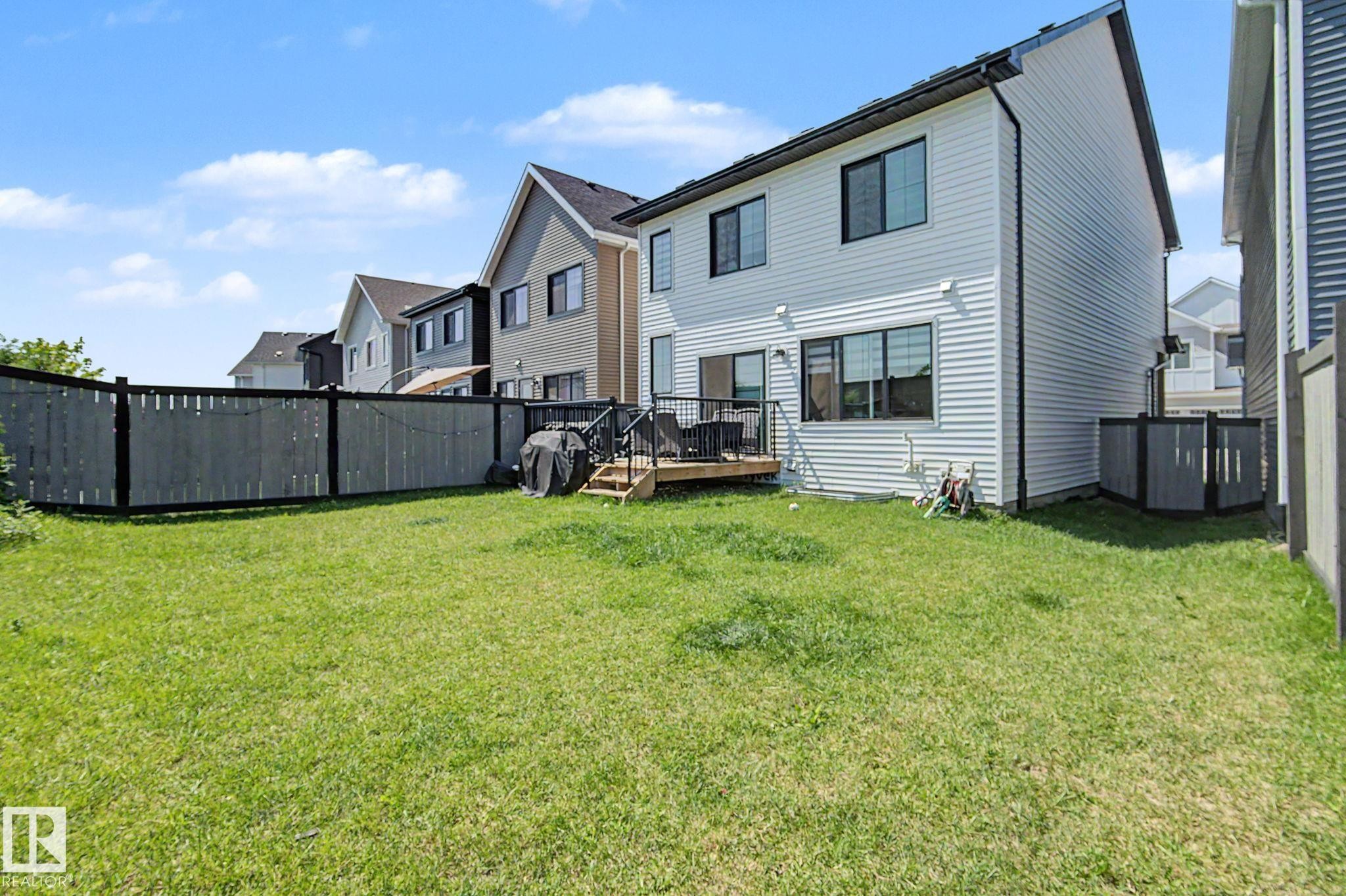 Expansive grassy yard featuring a wood privacy fence with black trim, a rear deck with dark railing, and a vinyl-sided exterior with black-framed windows - #12 905 172 St Sw, Edmonton, AB - Outdoor