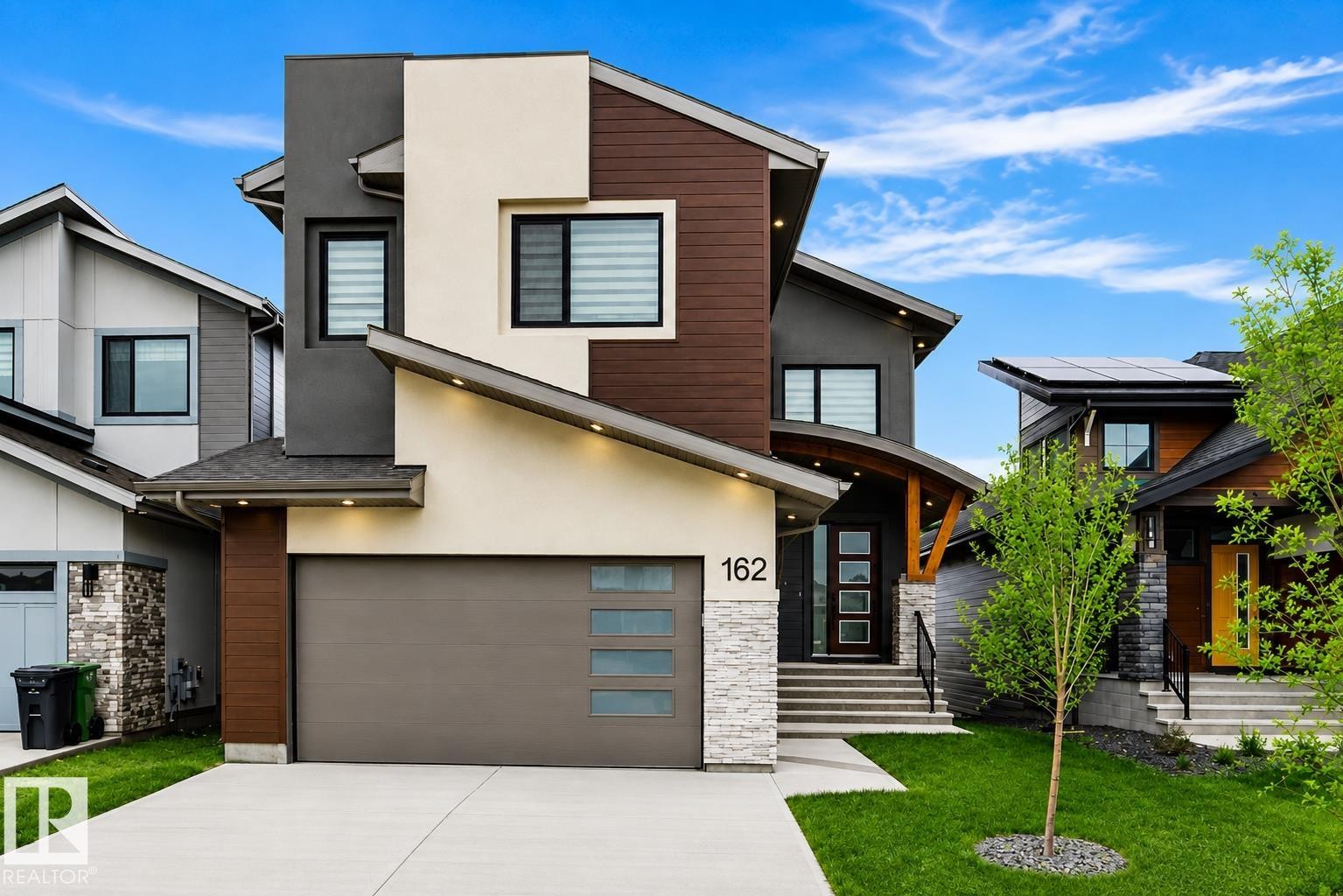 Contemporary exterior featuring multiple facade materials, an integrated garage, a covered entry with wood accents, and tiered front landscaping - 162 Edgewater Circle, Leduc, AB