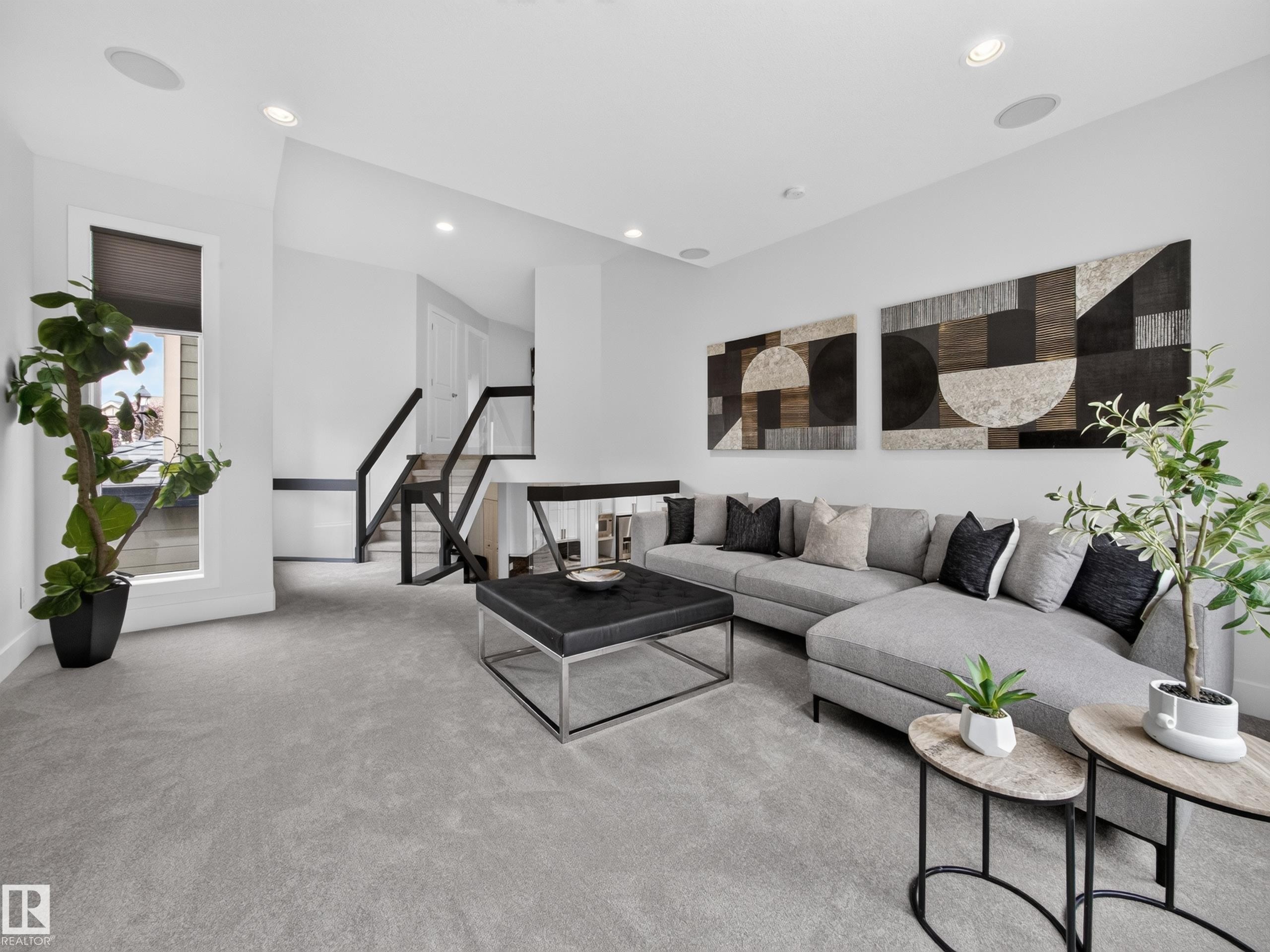 Spacious living area featuring light gray carpeting, recessed lighting, and a contemporary open-tread staircase with black railings - #59 10550 Ellerslie Rd Sw, Edmonton, AB - Indoor Photo Showing Living Room