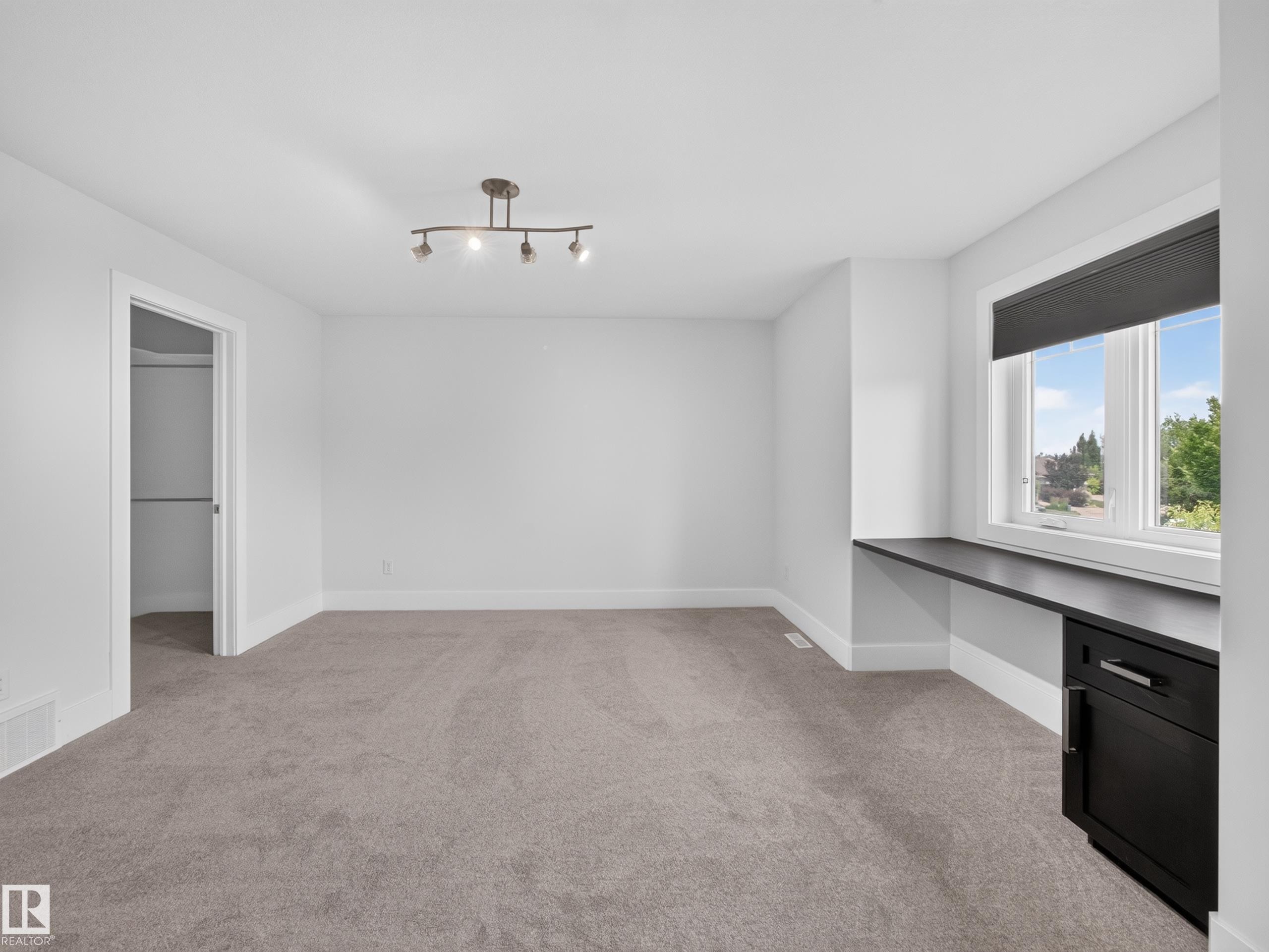 Spacious room featuring neutral carpeting, white walls, a built-in dark wood desk with a single cabinet, a large window, and a track lighting fixture - #59 10550 Ellerslie Rd Sw, Edmonton, AB - Indoor Photo Showing Other Room
