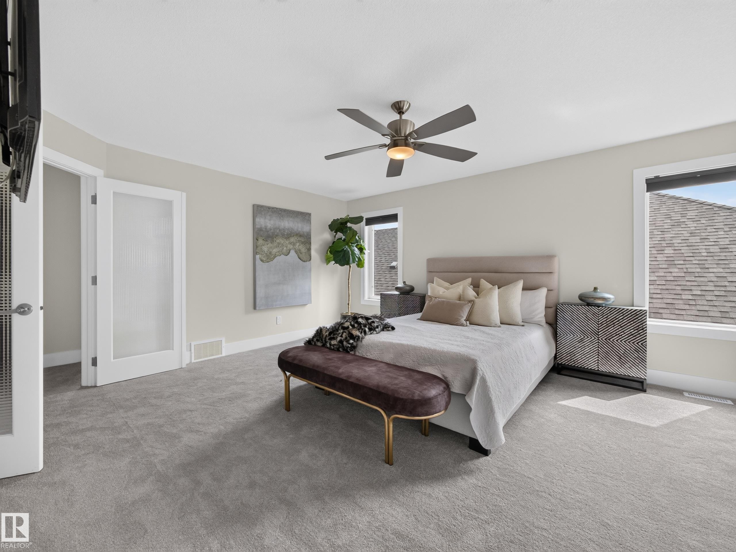 Carpeted bedroom featuring a ceiling fan with integrated lighting, two large windows, a frosted pane door, and neutral wall tones - #59 10550 Ellerslie Rd Sw, Edmonton, AB - Indoor Photo Showing Bedroom