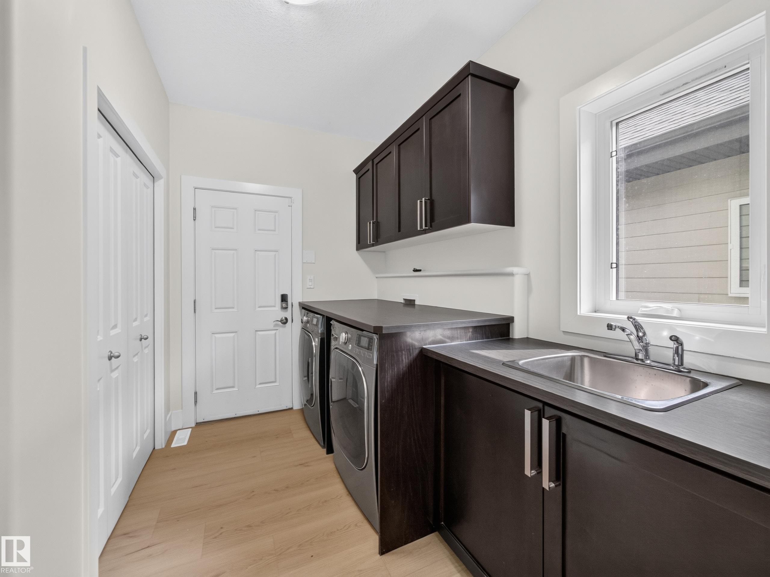 Laundry room featuring dark wood-finish cabinetry, a stainless steel utility sink, a large window, a light wood-finish floor, and white paneled doors - #59 10550 Ellerslie Rd Sw, Edmonton, AB - Indoor Photo Showing Laundry Room