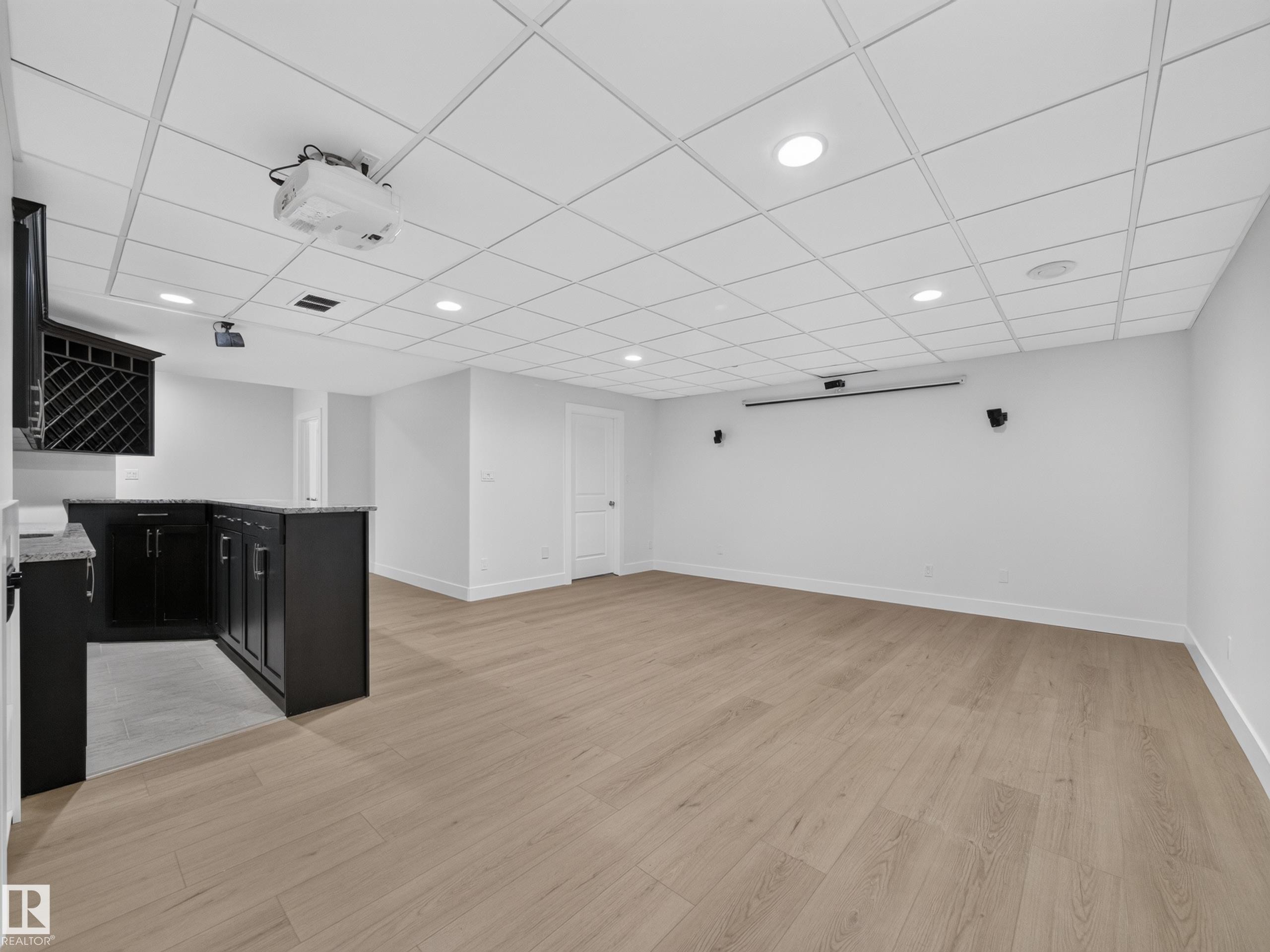 Finished basement featuring wood-finish flooring, a built-in wet bar with dark cabinetry and stone-look countertops, a drop ceiling with recessed lighting, a ceiling-mounted projector, and a retractable screen - #59 10550 Ellerslie Rd Sw, Edmonton, AB - Indoor Photo Showing Other Room