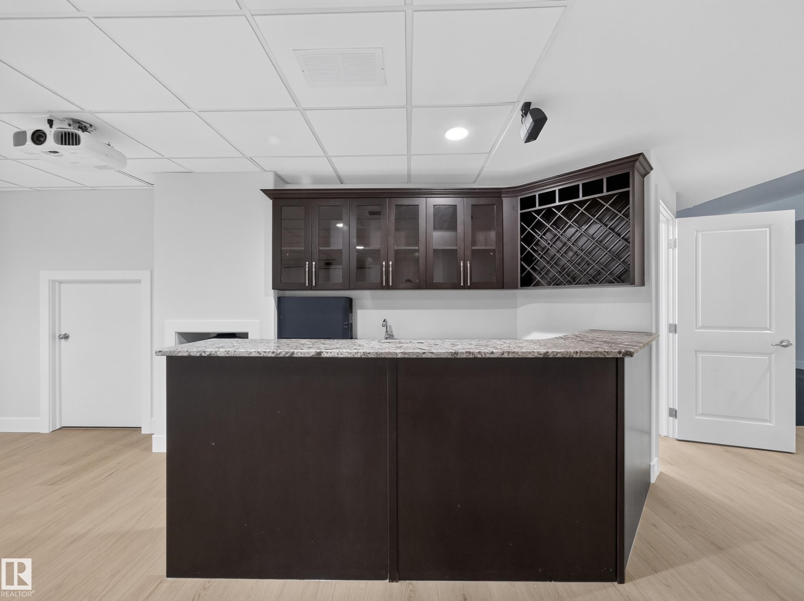 Wet bar featuring a light-toned countertop, dark wood-finish cabinetry, a built-in wine rack, and a sink with a faucet - #59 10550 Ellerslie Rd Sw, Edmonton, AB - Indoor