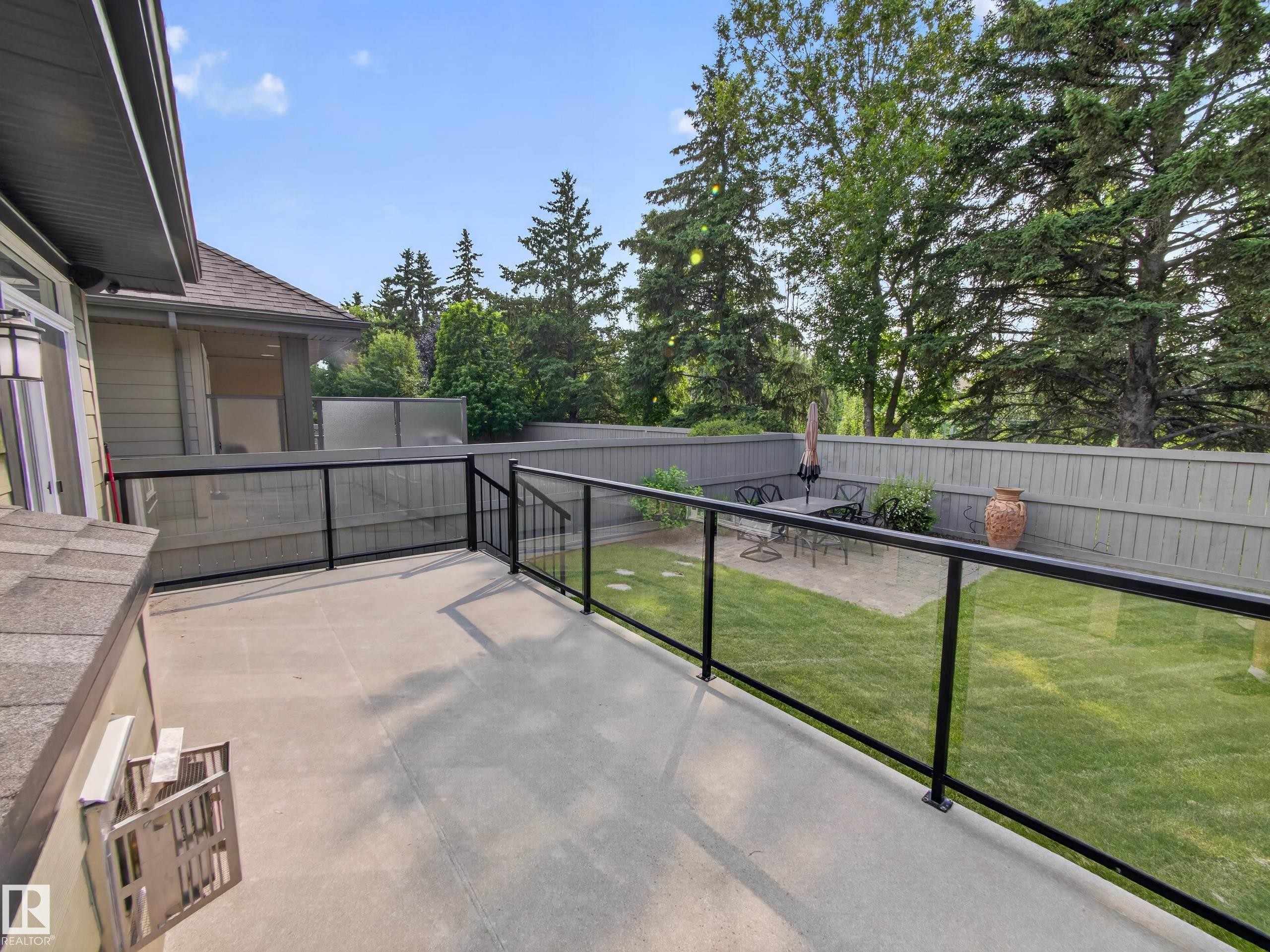 Spacious concrete deck featuring clear glass railings with black metal frames, a solid privacy fence, and a manicured lawn area with mature trees - #59 10550 Ellerslie Rd Sw, Edmonton, AB - Outdoor