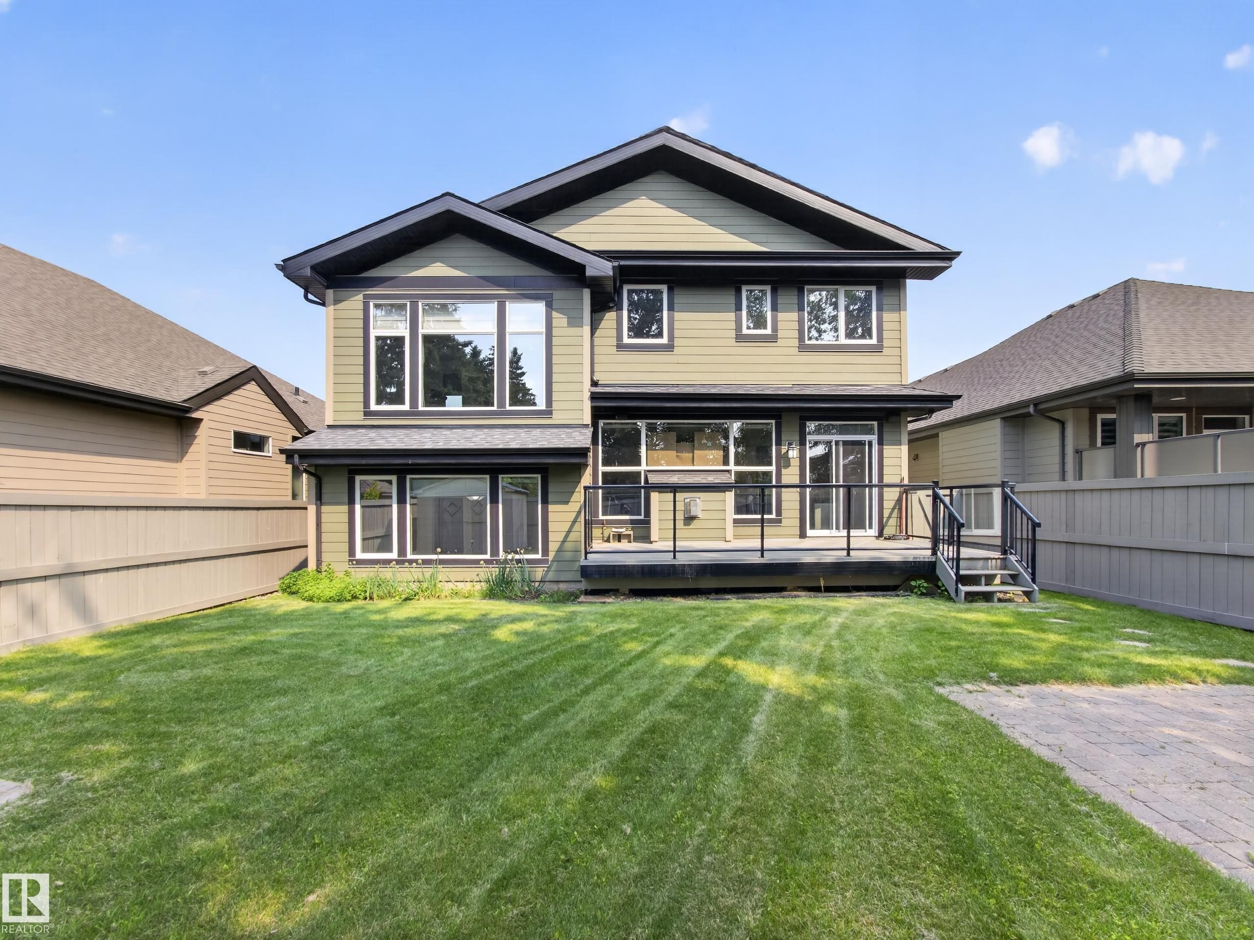 Rear exterior featuring an expansive grassy lawn, large windows, an upper-level balcony, a ground-level deck with black railings, and a side paver path - #59 10550 Ellerslie Rd Sw, Edmonton, AB - Outdoor