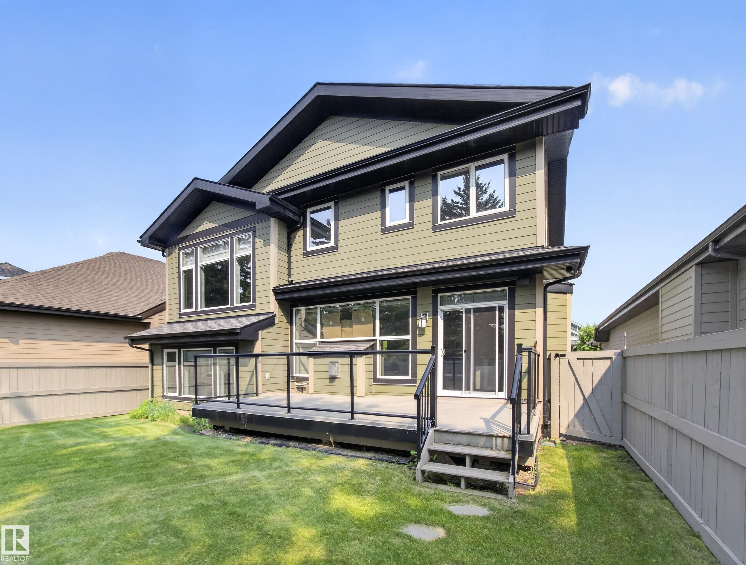 Contemporary residence featuring an olive green facade with dark trim, a spacious rear deck with glass railings, a sliding patio door, and a manicured lawn - #59 10550 Ellerslie Rd Sw, Edmonton, AB - Outdoor