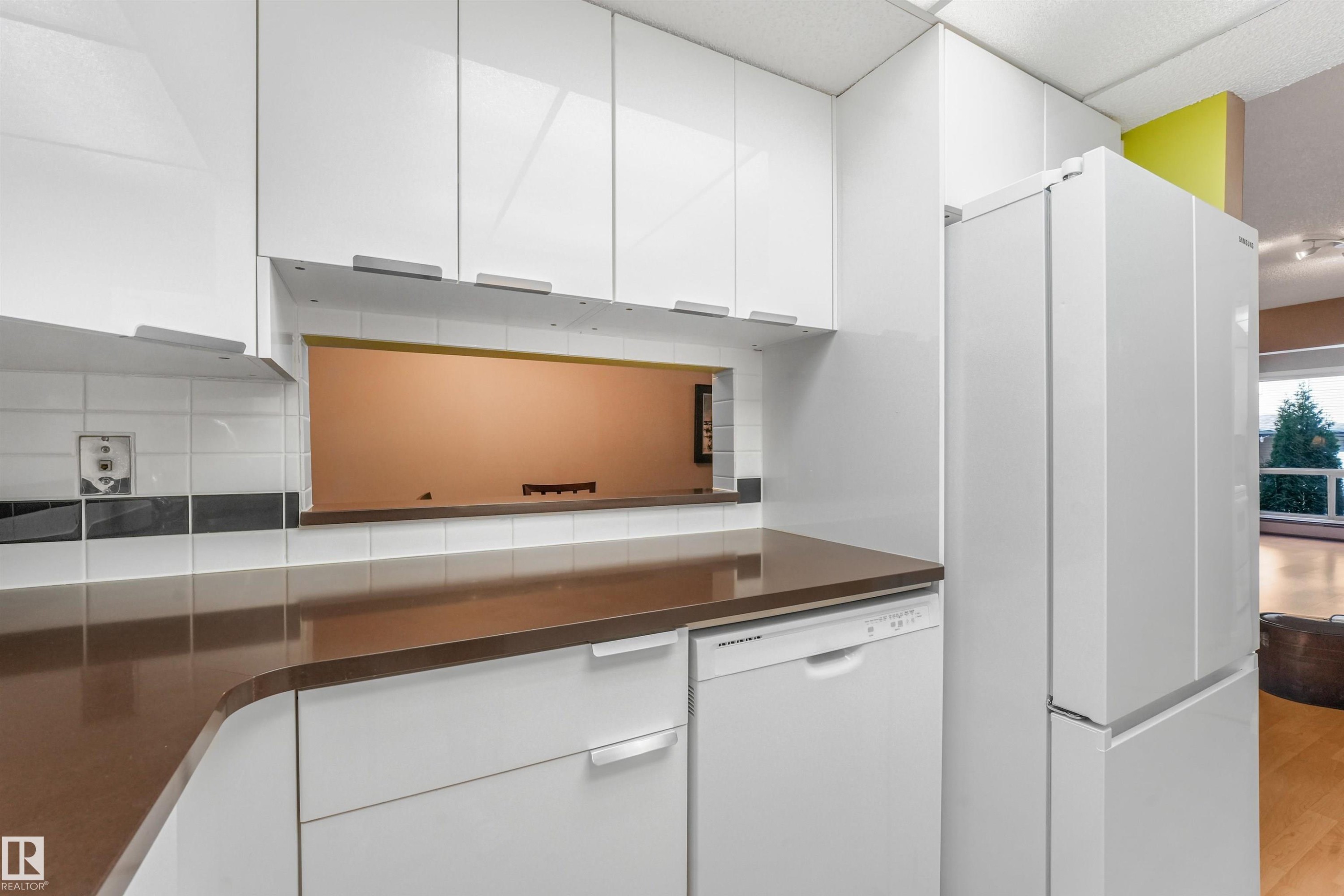 White high-gloss cabinetry, brown solid-surface countertops, integrated dishwasher, white subway tile backsplash, and a pass-through window - #203 10171 119 St Nw, Edmonton, AB