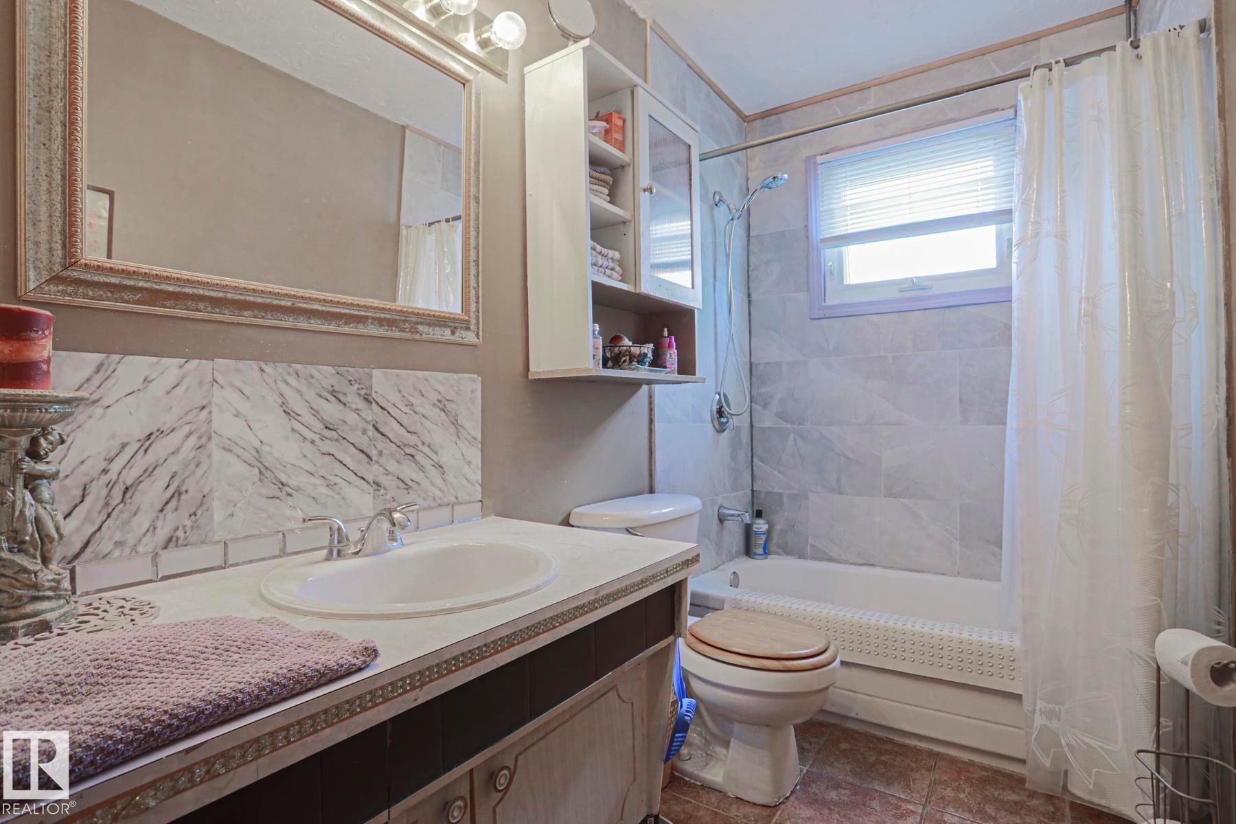 Bathroom featuring a vanity with an integrated sink, a framed wall mirror, a tiled shower/tub combination, wall-mounted cabinetry, and a window with blinds - 11440 144 Avenue Nw, Edmonton, AB