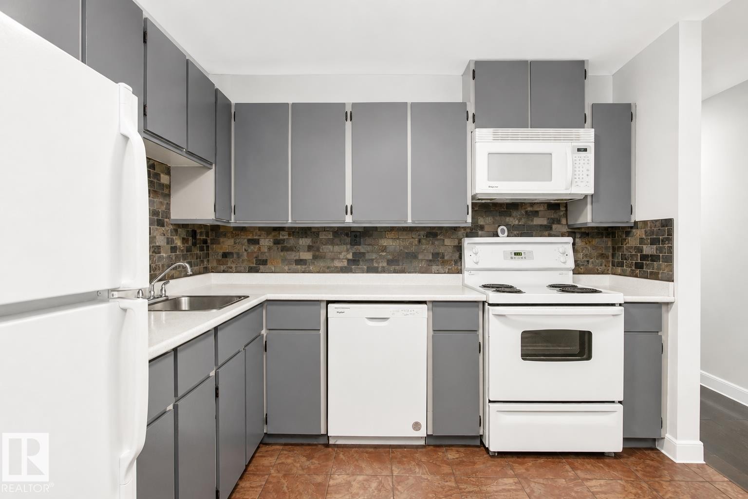 Efficient galley kitchen featuring gray cabinetry with chrome hardware, white appliances including electric range and microwave, tile backsplash, laminate countertops, and wood-finish flooring throughout - #203 2624 Mill Woods Rd E Nw, Edmonton, AB