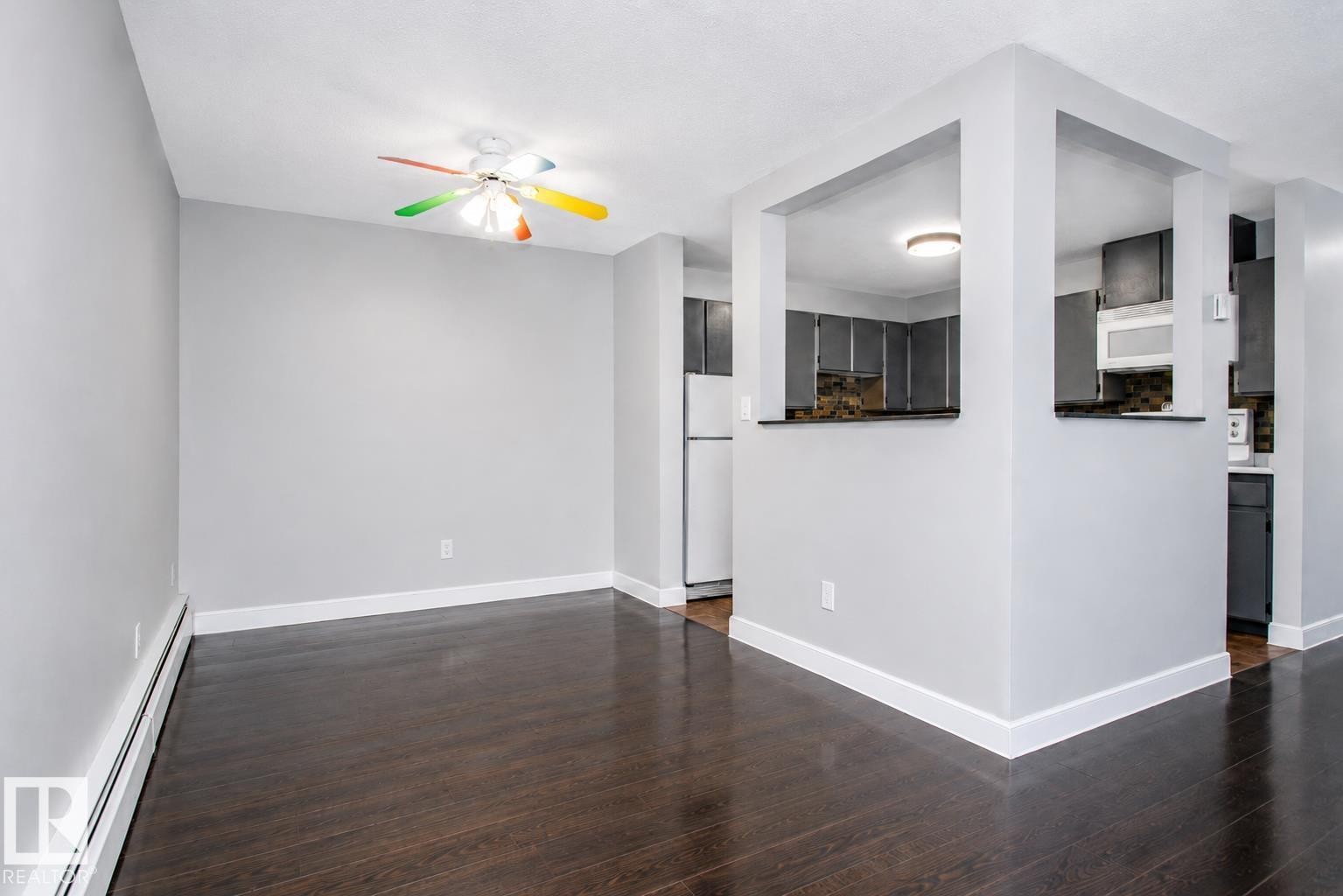 Wood-finish flooring throughout, light gray wall color, white ceiling, ceiling fan with multicolored blades, and a pass-through opening to the kitchen featuring dark cabinetry - #203 2624 Mill Woods Rd E Nw, Edmonton, AB