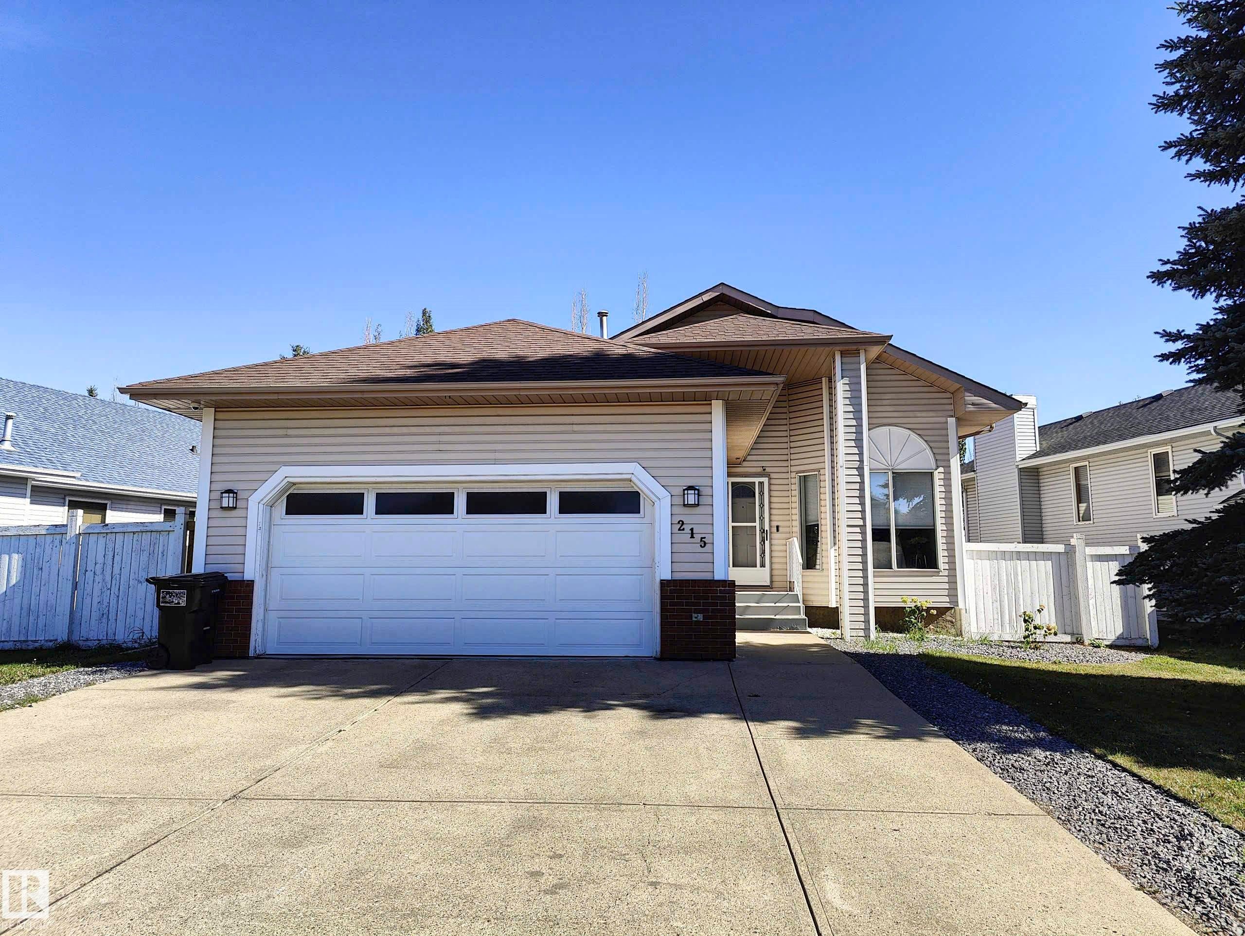 Front exterior featuring a concrete driveway, attached garage with panel doors, and a partial white privacy fence - 215 Peter Close Nw, Edmonton, AB