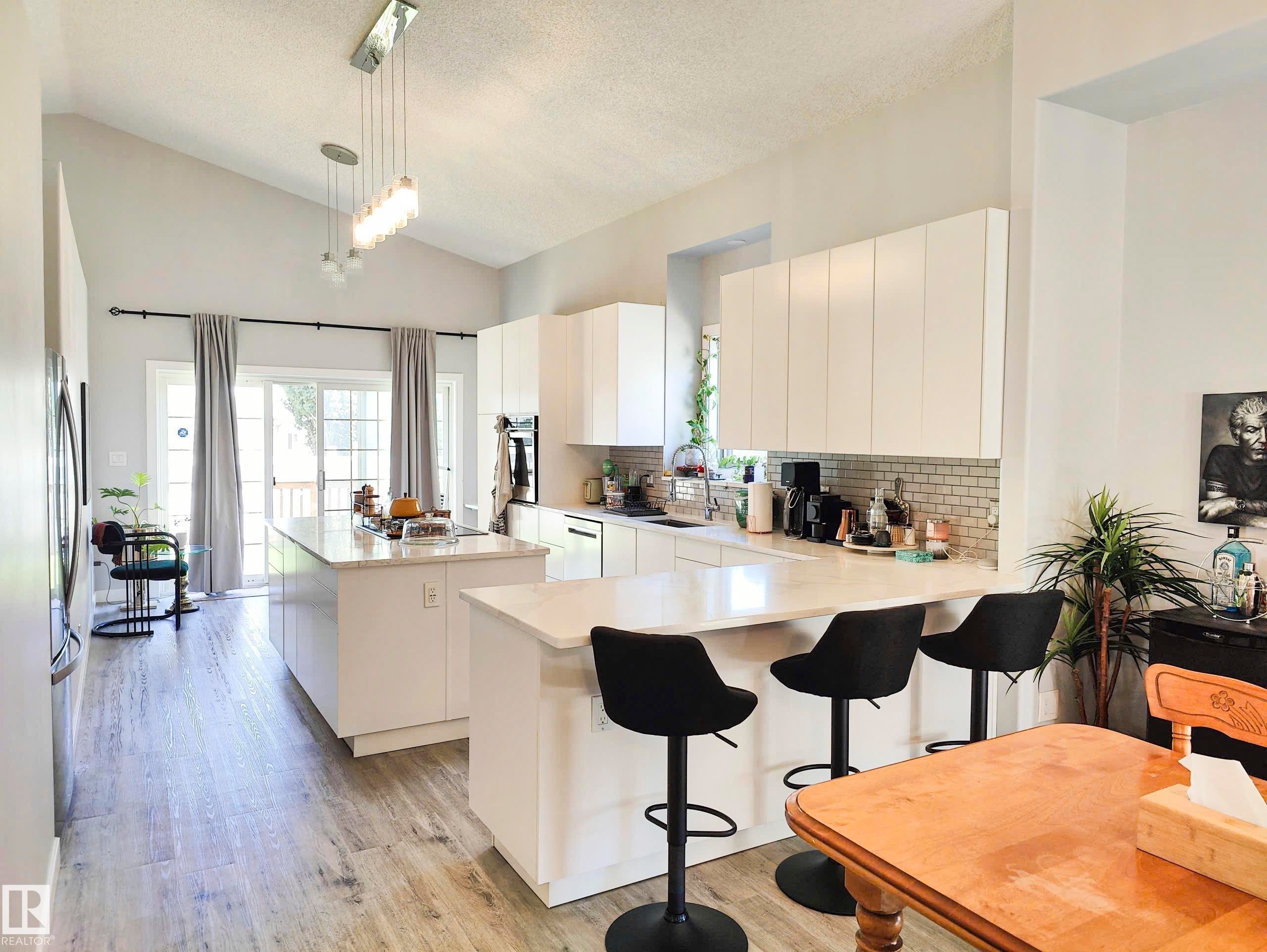 Vaulted ceiling with an architectural chandelier, wood-finish flooring, two kitchen islands with solid surface countertops, white flat-panel cabinetry, and a subway tile backsplash - 215 Peter Close Nw, Edmonton, AB