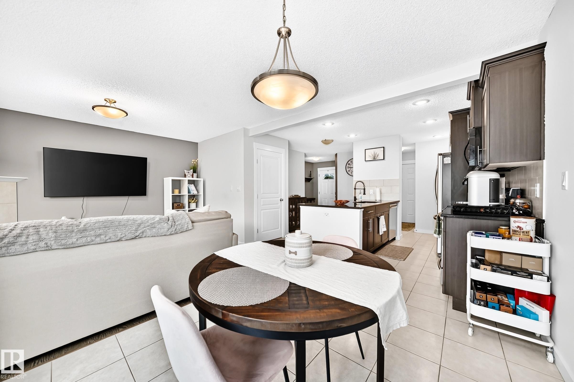 Open-concept living space featuring a dining area with a wood-finish table, a kitchen with dark cabinetry and stainless steel appliances, and tile flooring throughout - 16832 53 Street Nw, Edmonton, AB