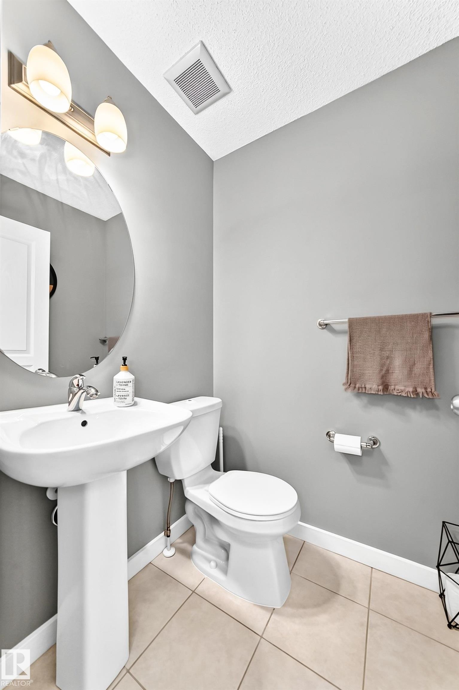 Pedestal sink with chrome faucet, round wall-mounted mirror, ceramic tile flooring, and integrated ceiling ventilation - 16832 53 Street Nw, Edmonton, AB