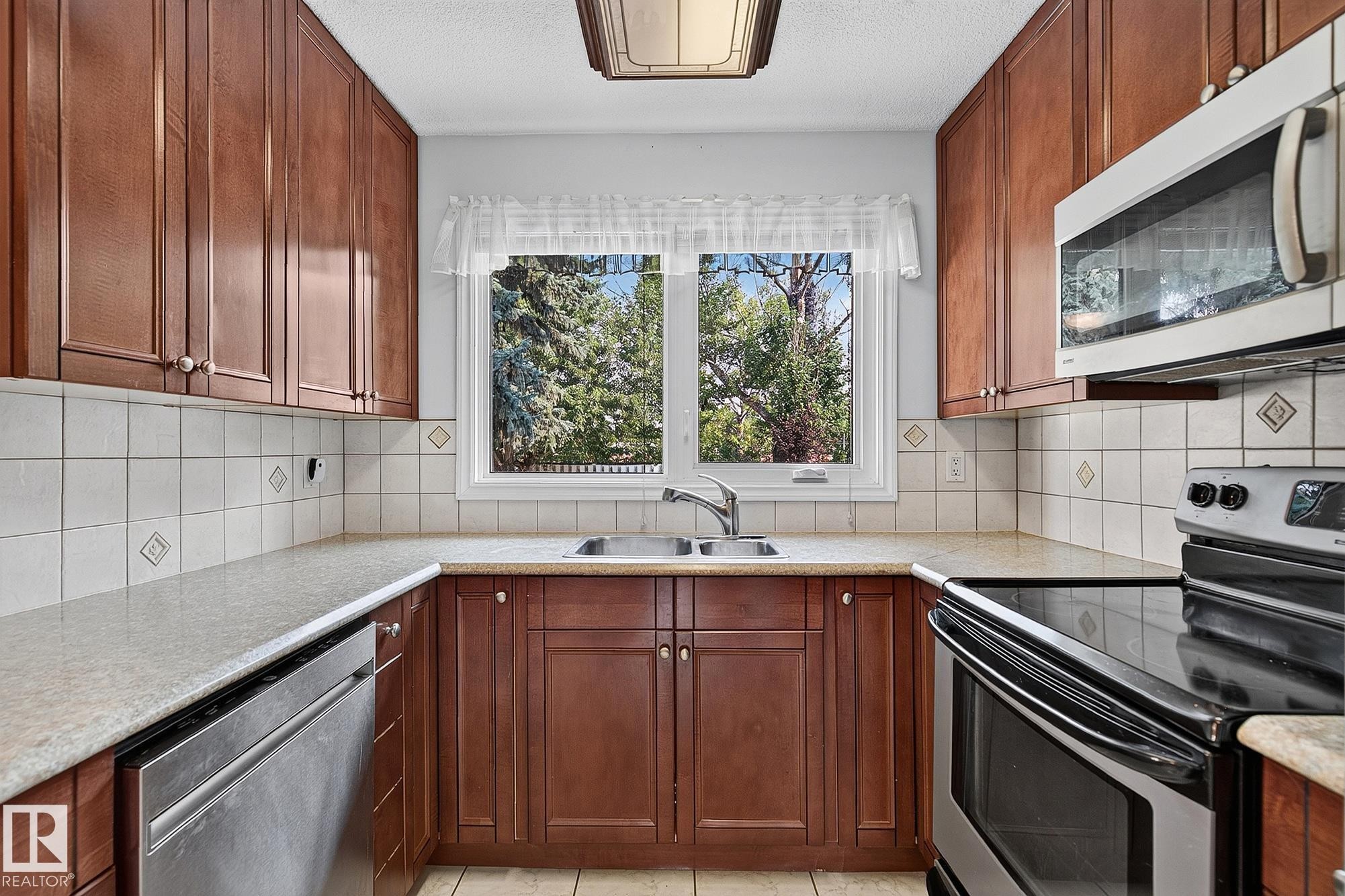 Kitchen featuring wood cabinetry, white tile backsplash, light-toned countertops, a stainless steel sink, and stainless steel appliances - 16016 78A Avenue Nw, Edmonton, AB