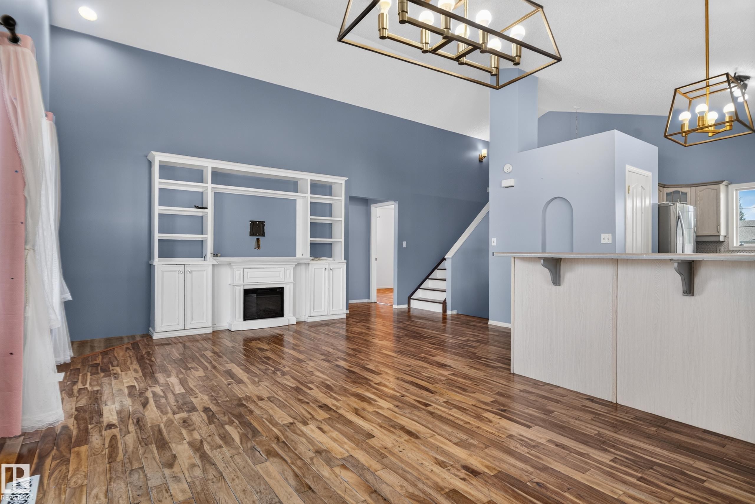 Vaulted ceilings with a decorative chandelier, wood-finish flooring, built-in entertainment center with a fireplace, and a kitchen island with a counter overhang - 13803 130A Avenue Nw, Edmonton, AB