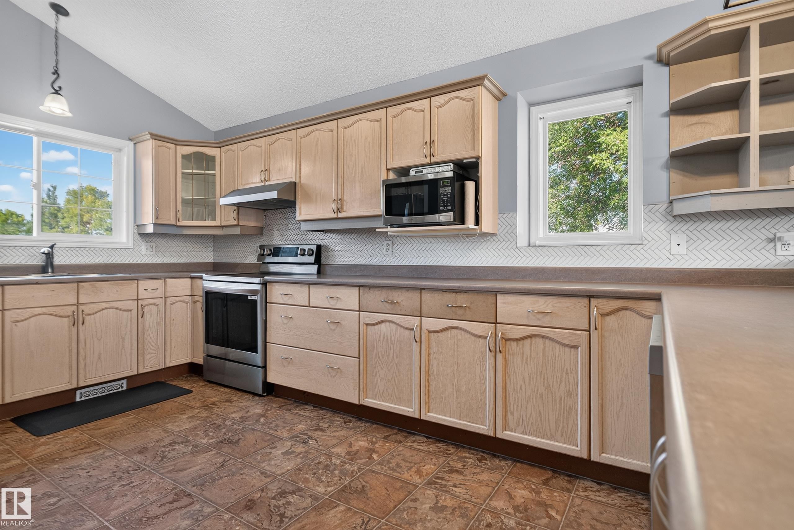 Kitchen featuring extensive wood-finish cabinetry, stainless steel appliances, a herringbone tile backsplash, and a vaulted ceiling - 13803 130A Avenue Nw, Edmonton, AB