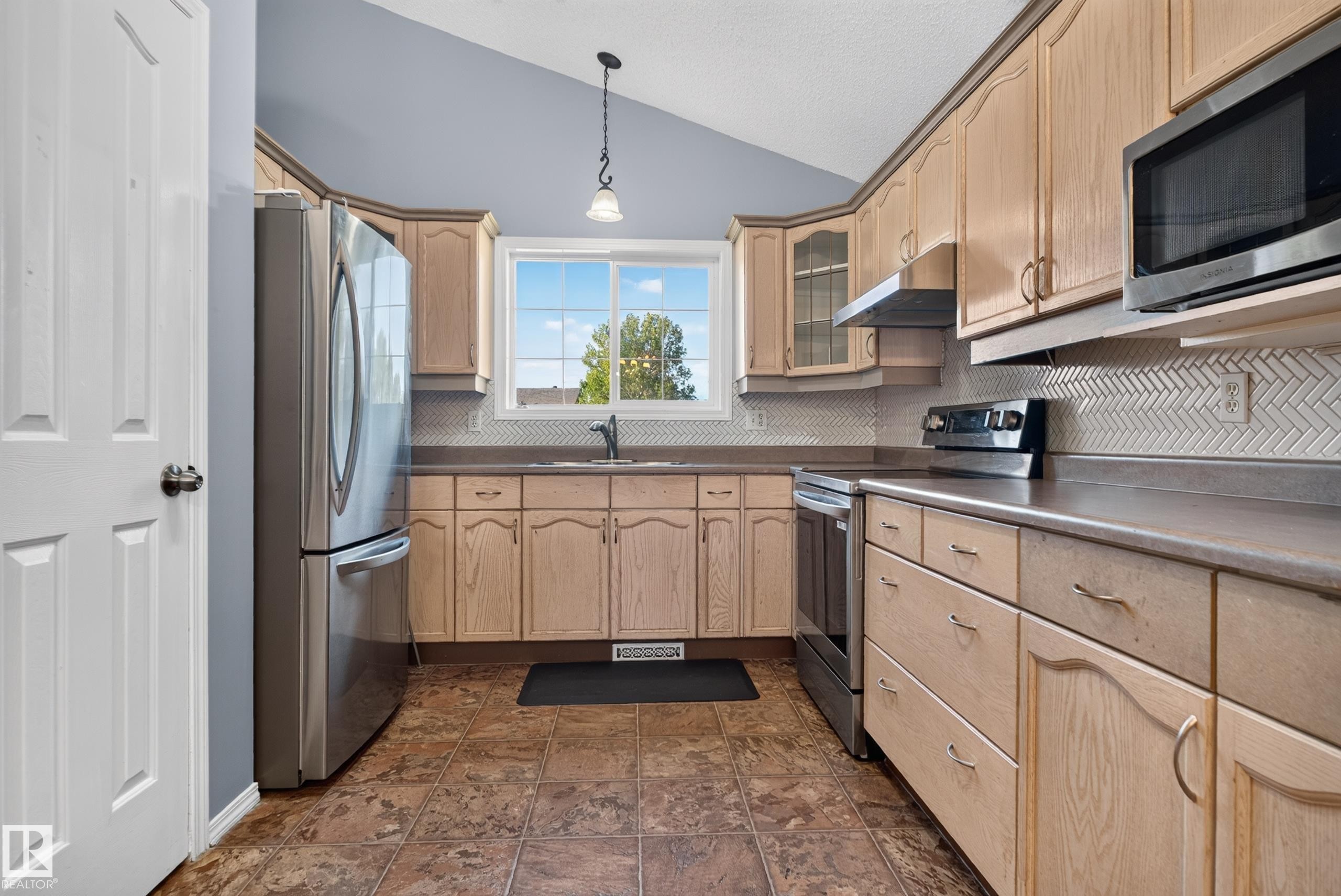 Kitchen featuring a vaulted ceiling, wood-finish cabinetry, stainless steel appliances, herringbone tile backsplash, and a window above the sink - 13803 130A Avenue Nw, Edmonton, AB
