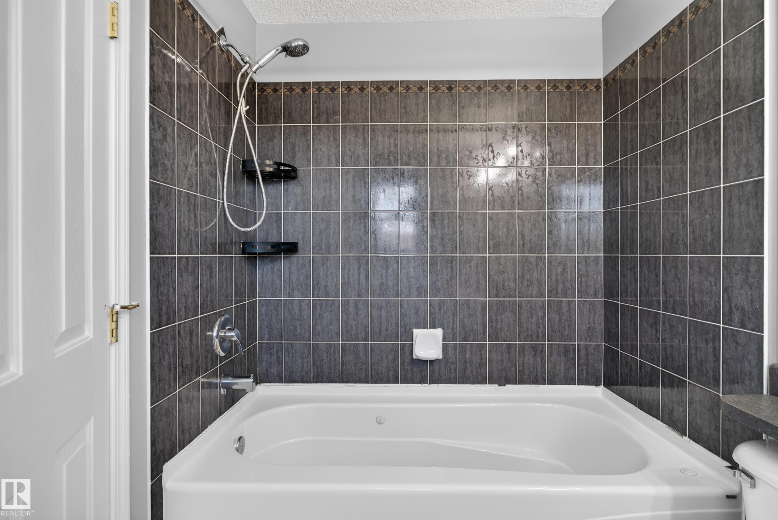 White soaking tub with integrated jets, dark gray tile surround, decorative tile accent band, and chrome shower fixtures - 13803 130A Avenue Nw, Edmonton, AB