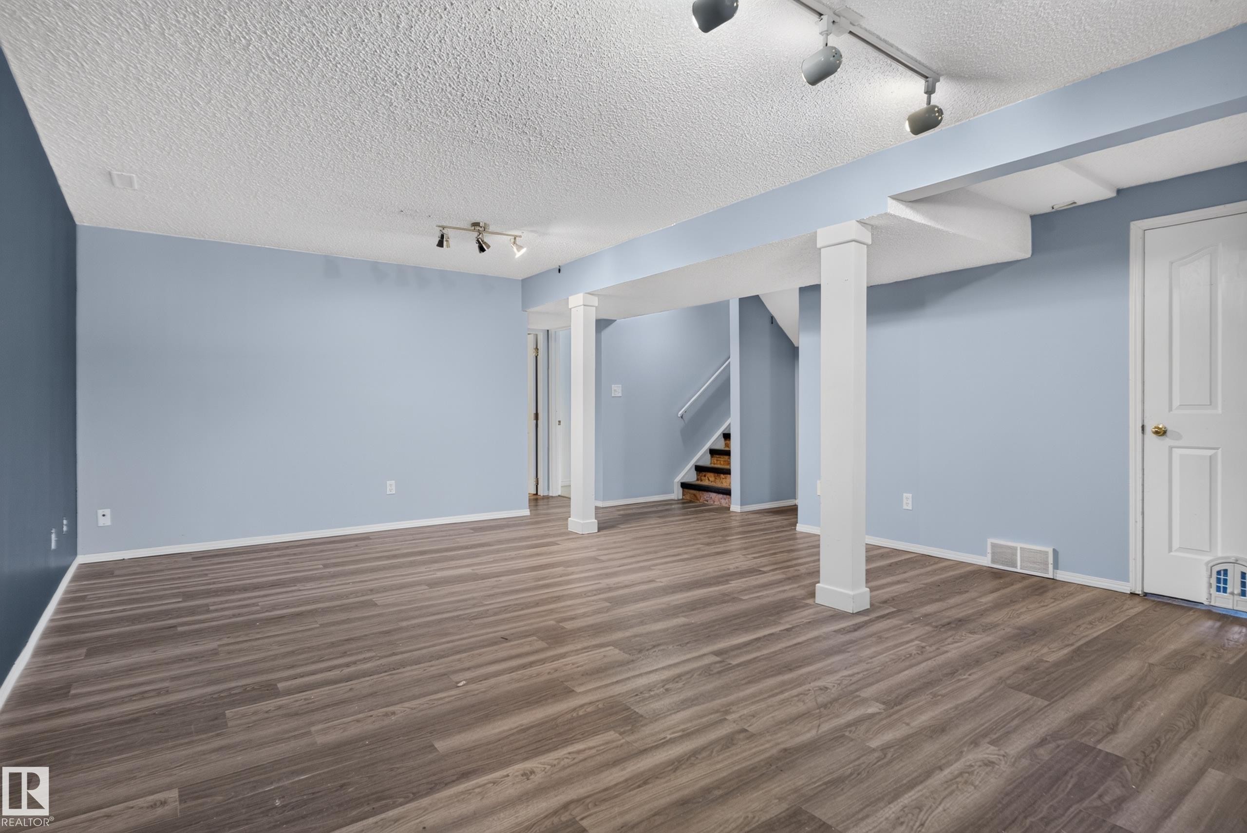 Expansive room featuring wood-finish flooring, light blue walls, exposed structural beams, white trim, and track lighting - 13803 130A Avenue Nw, Edmonton, AB