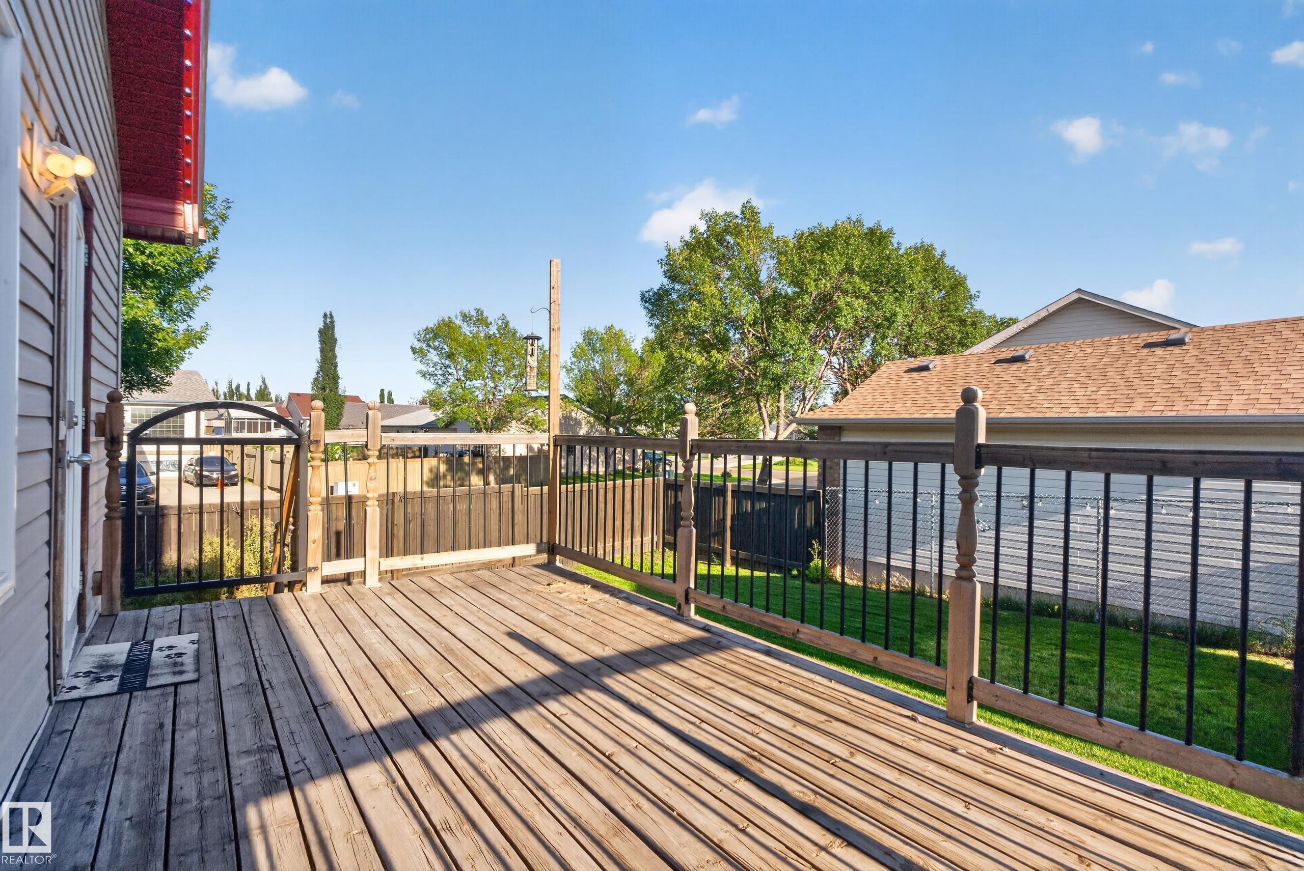 Wood-finish deck with a black metal and wood railing, featuring a black metal gate and a light fixture on the exterior wall - 13803 130A Avenue Nw, Edmonton, AB