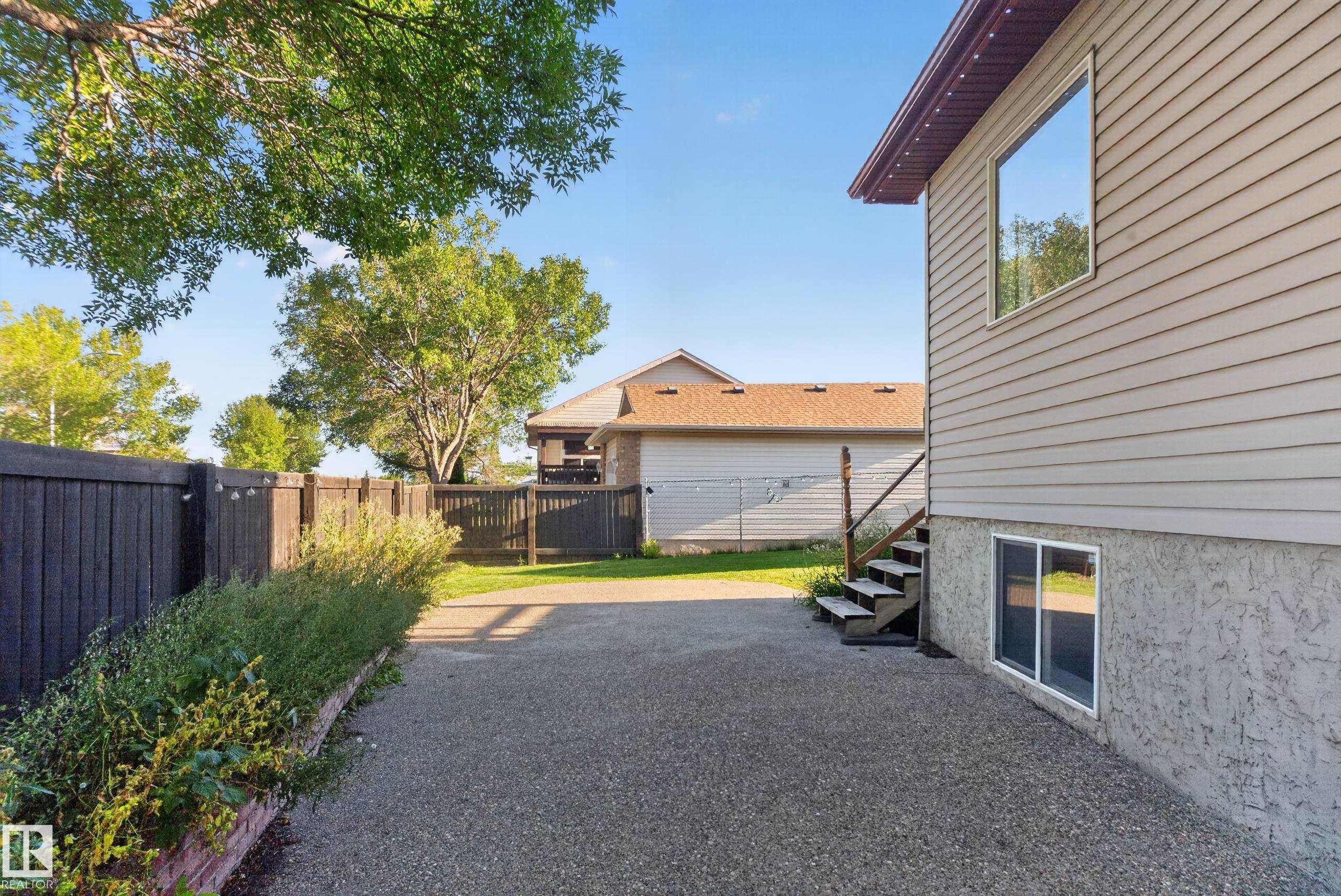Siding exterior with multiple windows, leading to a fenced yard with mature trees and a gravel surface - 13803 130A Avenue Nw, Edmonton, AB