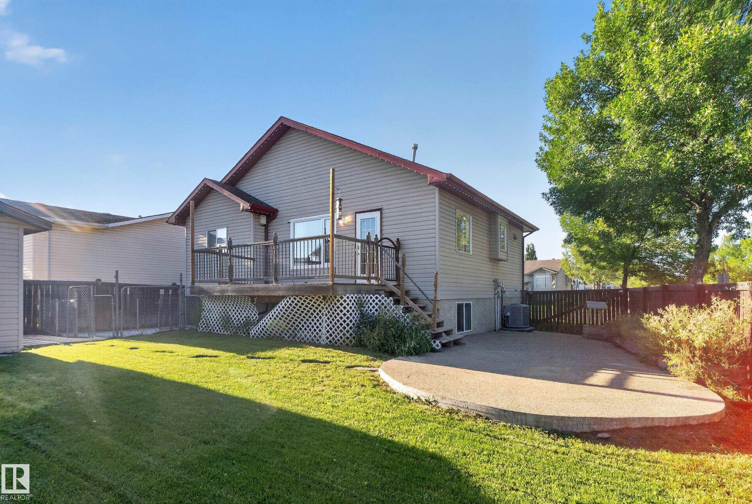 Elevated wood deck with lattice skirting, expansive green lawn, mature trees, and a curved concrete patio - 13803 130A Avenue Nw, Edmonton, AB