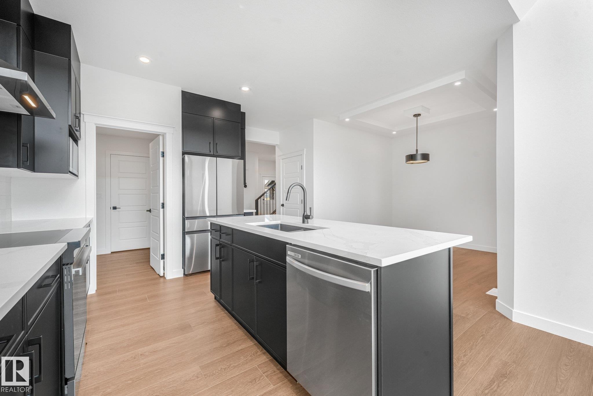 Contemporary kitchen featuring dark flat-panel cabinetry, stainless steel appliances, a center island with sink, light wood-finish flooring, and recessed lighting - 511 157 Avenue Ne, Edmonton, AB